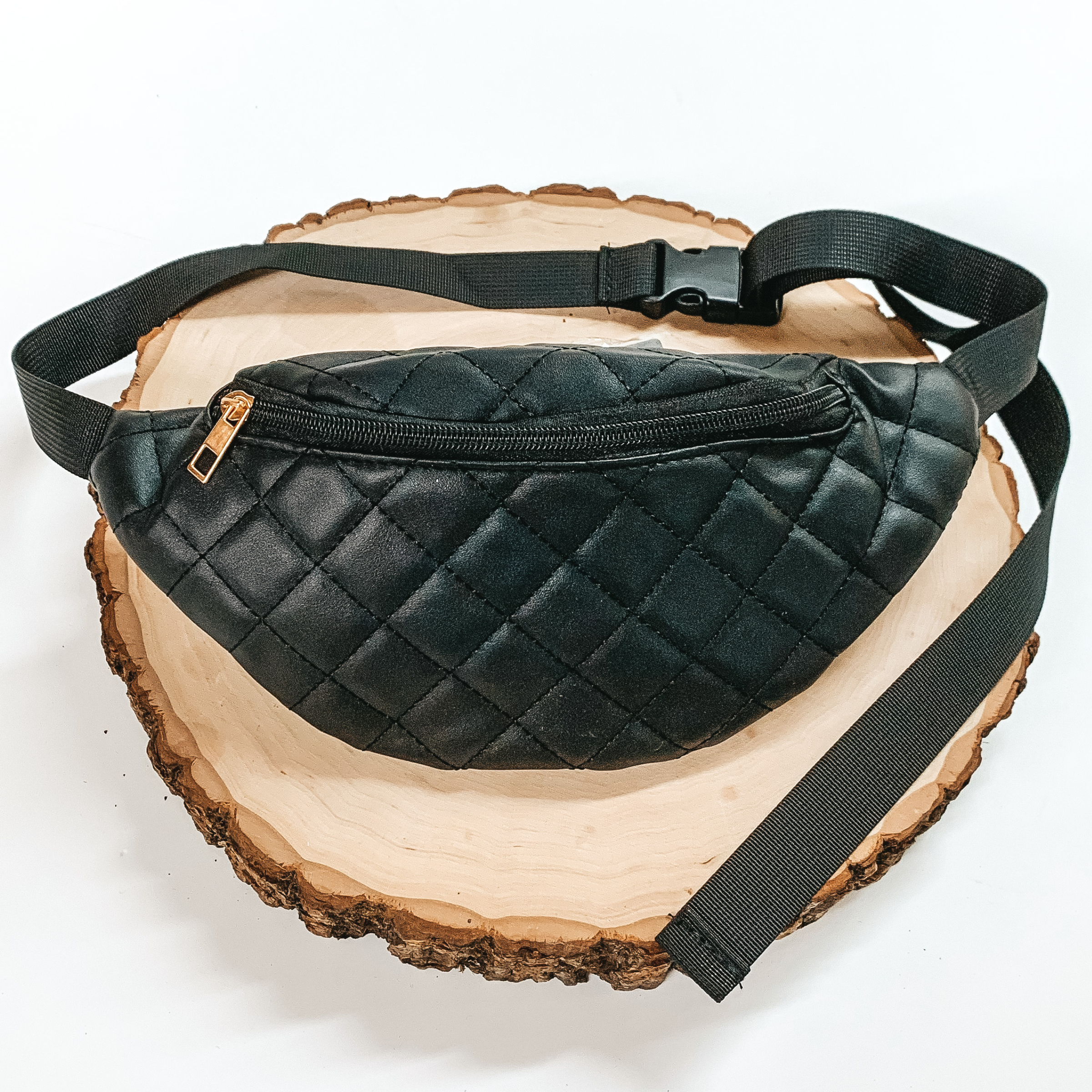 Black, quilted fanny pack with a zipper across the front. This fanny pack also includes black straps and black clips. This fanny pack is pictured on a piece of wood on a white background. 