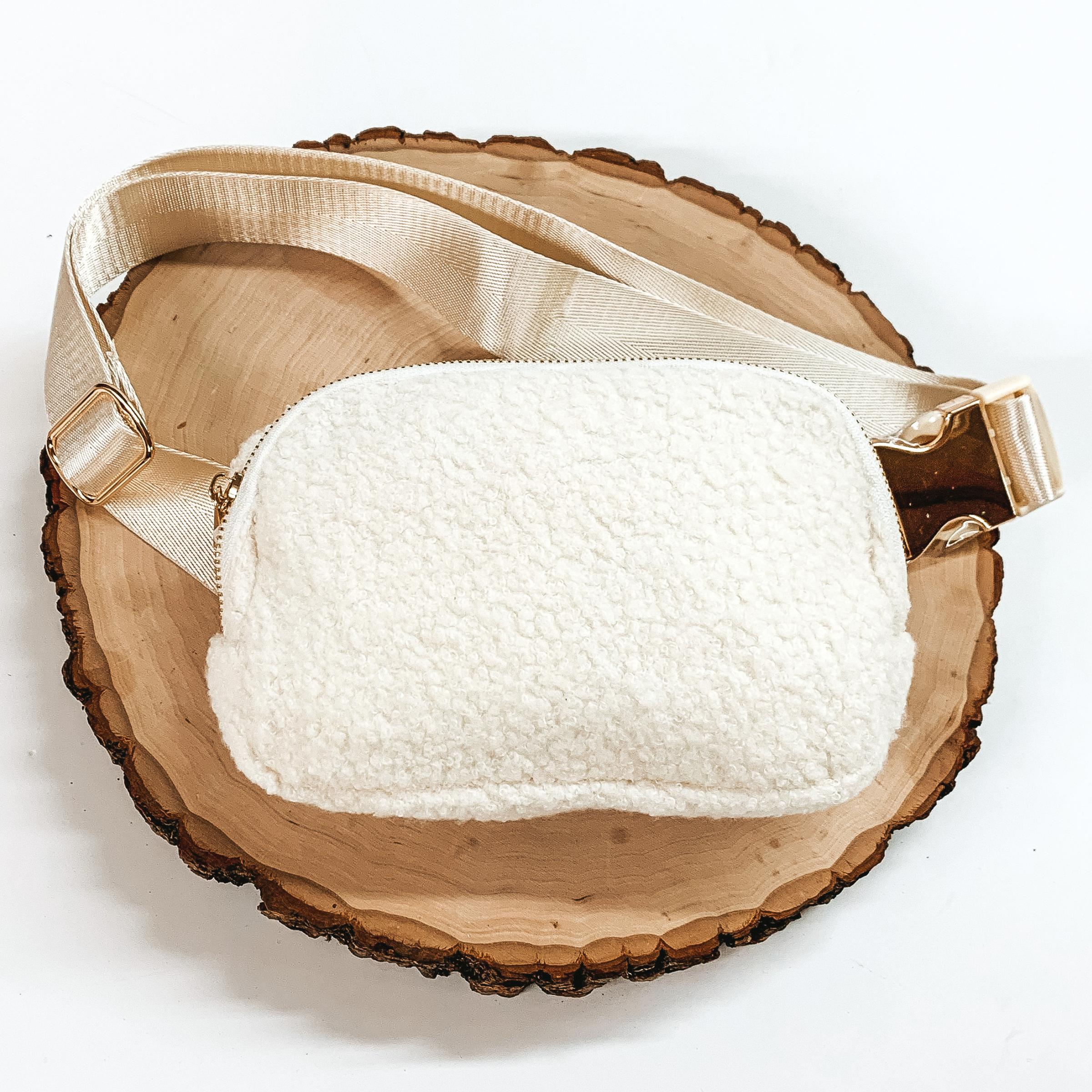 Ivory, sherpa fanny pack with a top zipper across the front. This fanny pack also includes ivory straps and gold clips. This fanny pack is pictured on a piece of wood on a white background. 