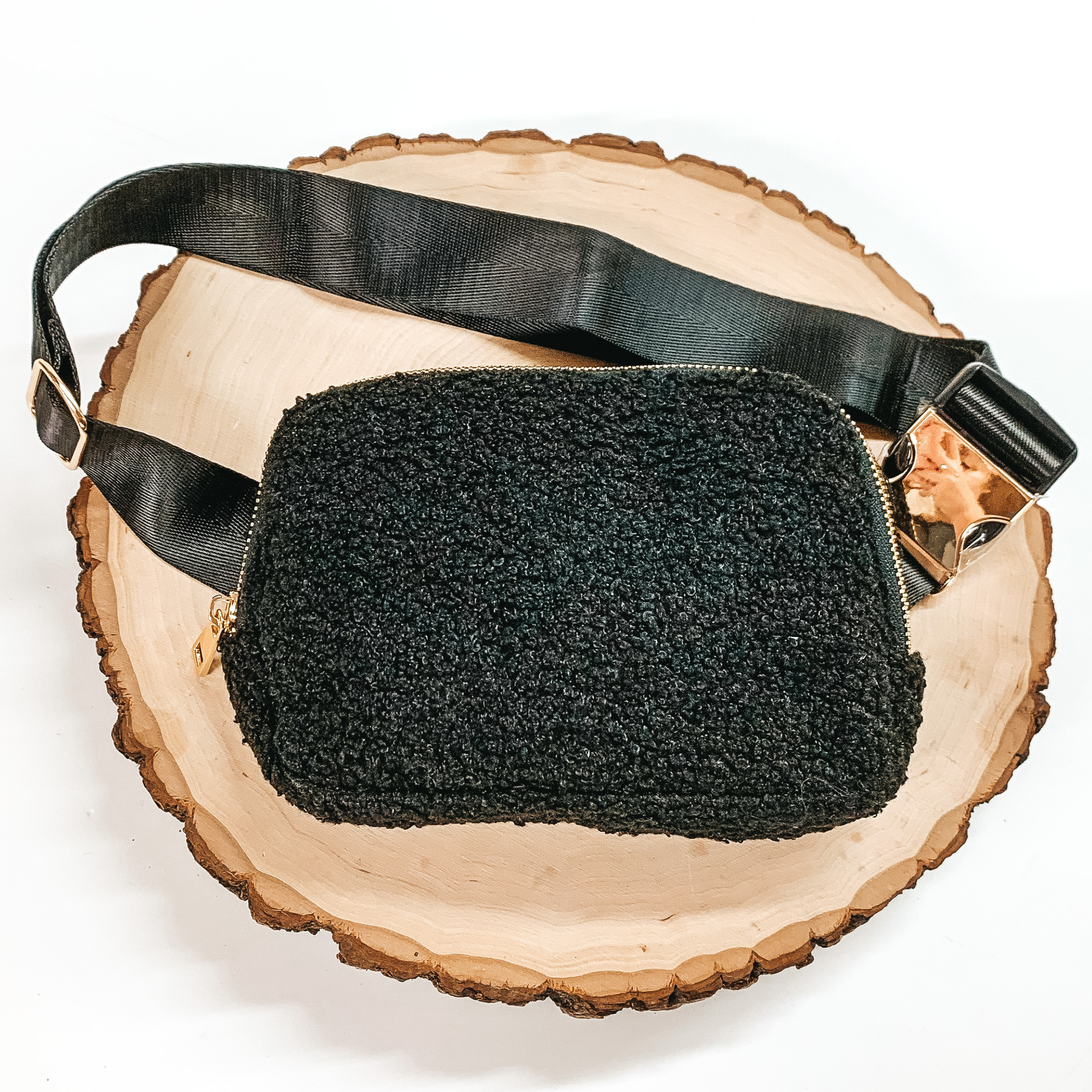 Black, sherpa fanny pack with a top zipper across the front. This fanny pack also includes black straps and gold clips. This fanny pack is pictured on a piece of wood on a white background. 