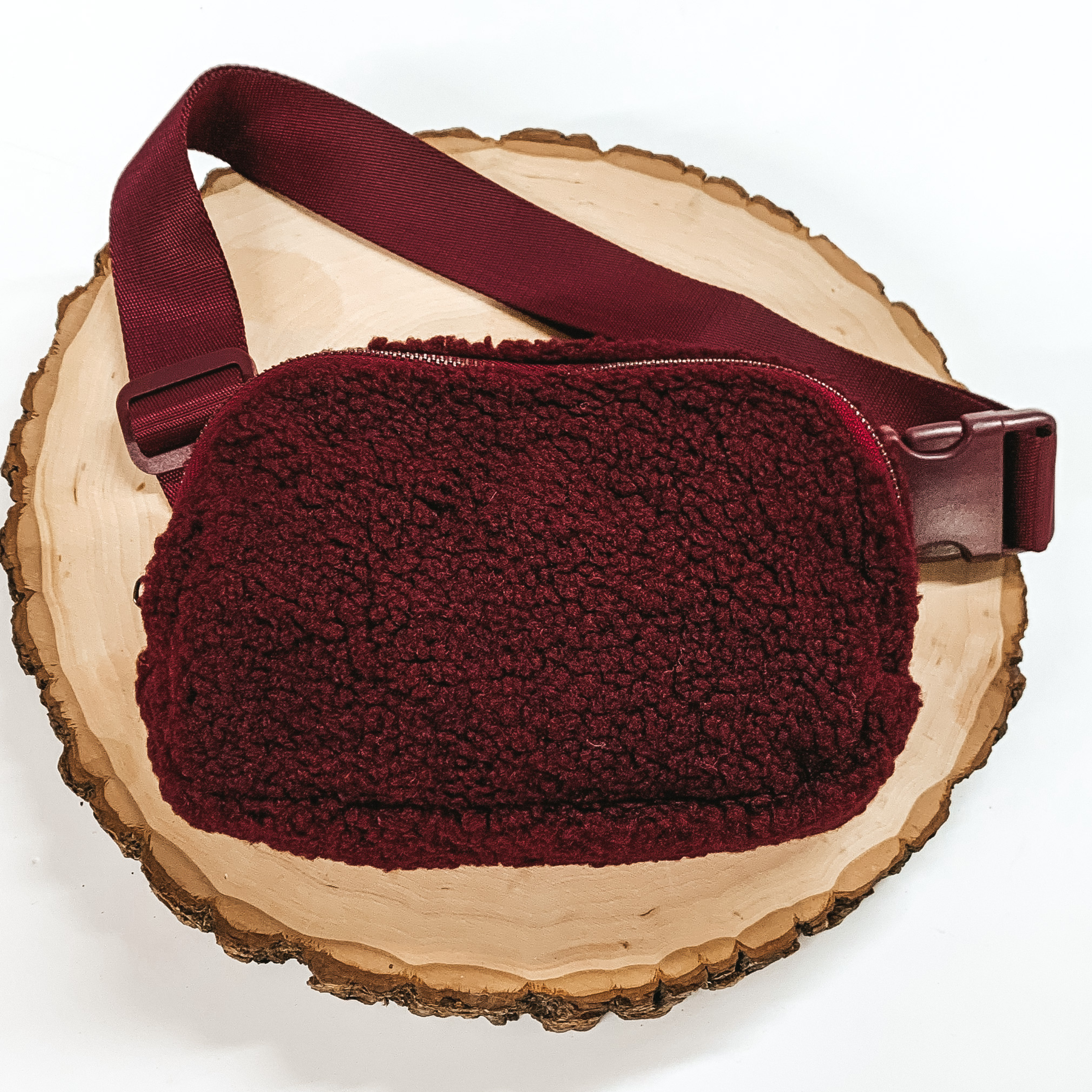 Maroon, sherpa fanny pack with a top zipper across the front. This fanny pack also includes maroon straps and maroon clips. This fanny pack is pictured on a piece of wood on a white background.
