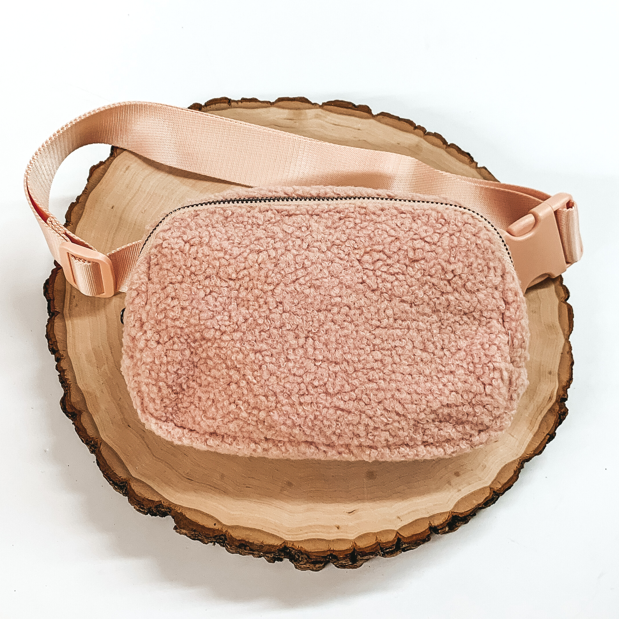 Blush pink, sherpa fanny pack with a top zipper across the front. This fanny pack also includes blush pink straps and blush pink clips. This fanny pack is pictured on a piece of wood on a white background. 