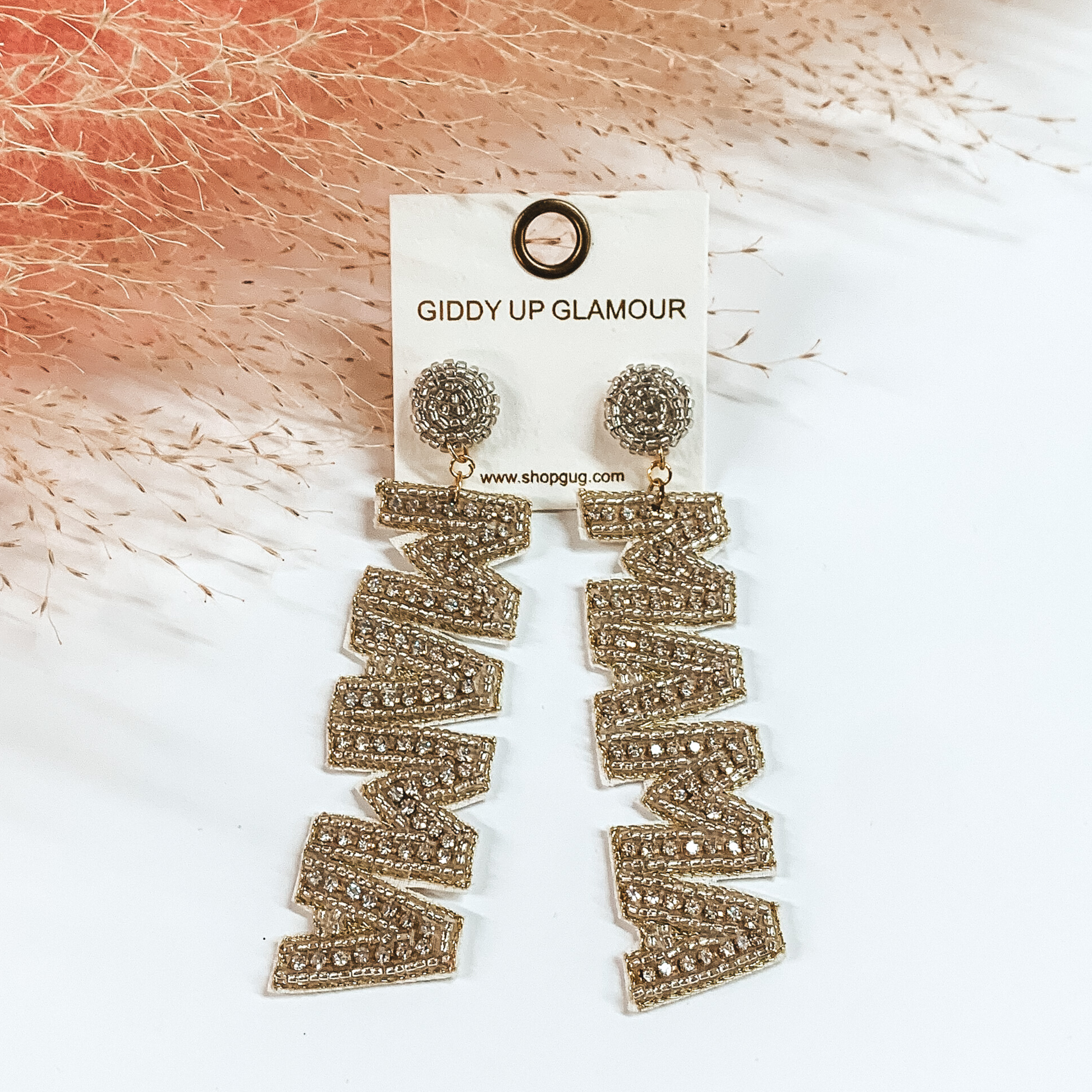 These earrings include silver, circle, beaded post back earrings with a long dangle. This dangle spells out "MAMA" in silver beads with an inlay of clear crystals. These earrings are pictured on a white background with pink pompous grass at the top. 