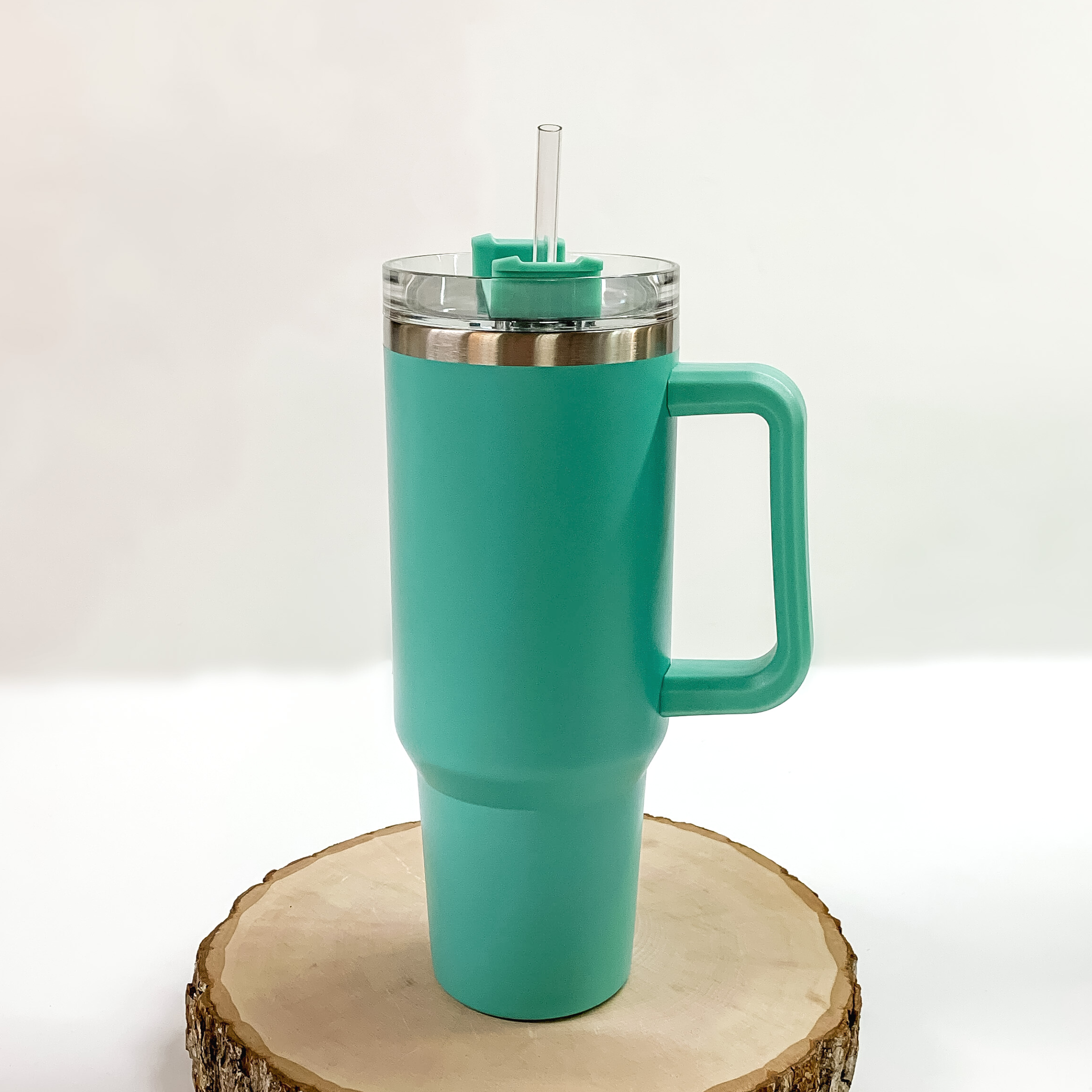 This is a mint tumbler with a mint handle. This tumbler also has a clear lid and straw. This tumbler is pictured standing on a piece of wood on a white background. 