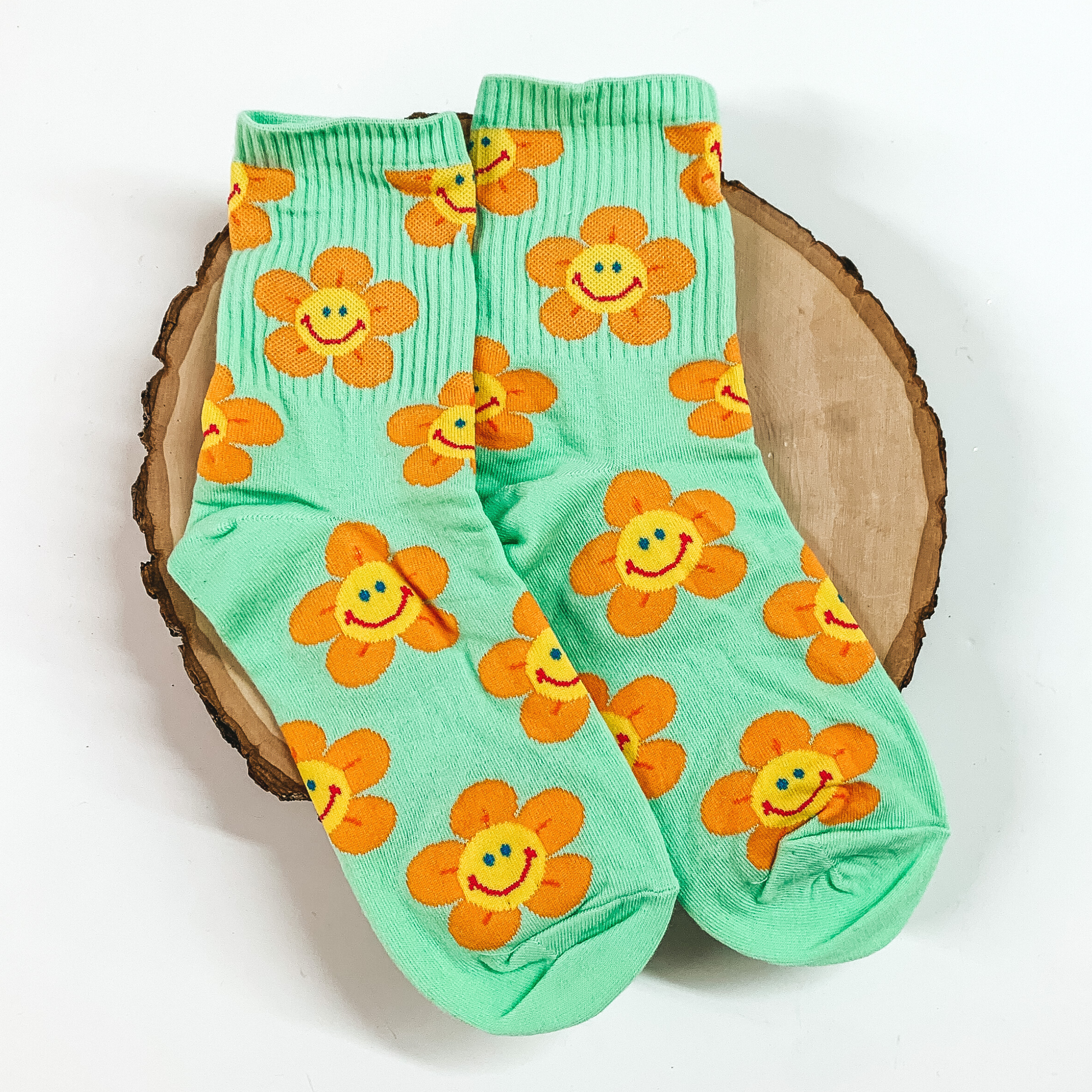 Mint ankle socks with an orange flower design. The flowers include a smiley face in the center. These socks are pictured on a piece of wood on a white background. 