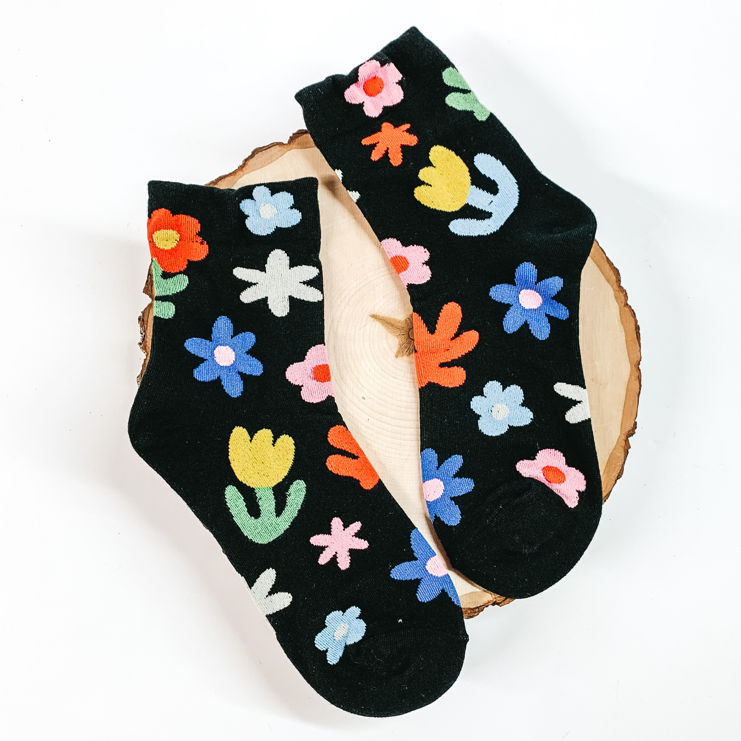 Black ankle socks that have a groovy flower design. The flower design is multicolored. These socks are pictured on a piece of wood on a white background. 
