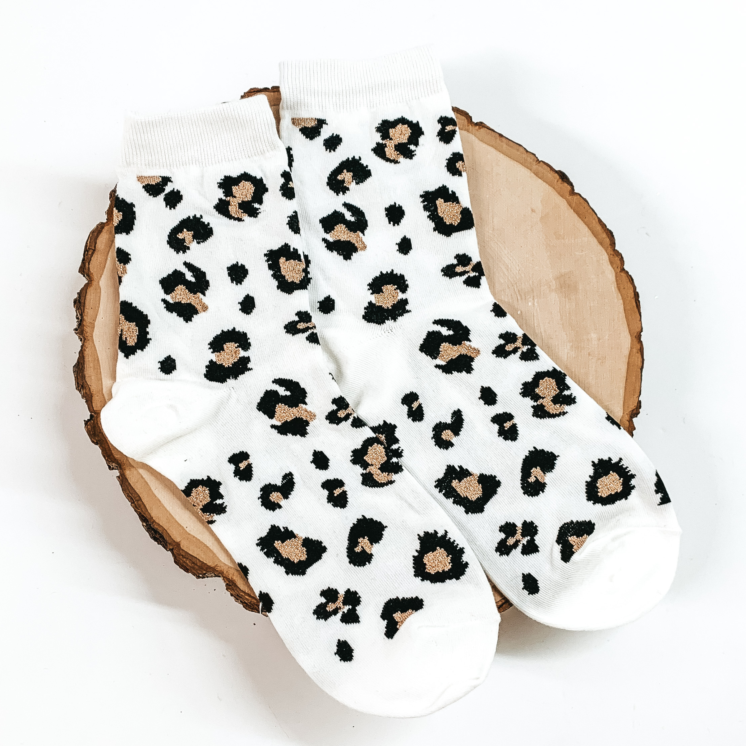 White ankle socks with a black and champagne leopard print design. These socks are pictured on a piece of wood on a white background. 