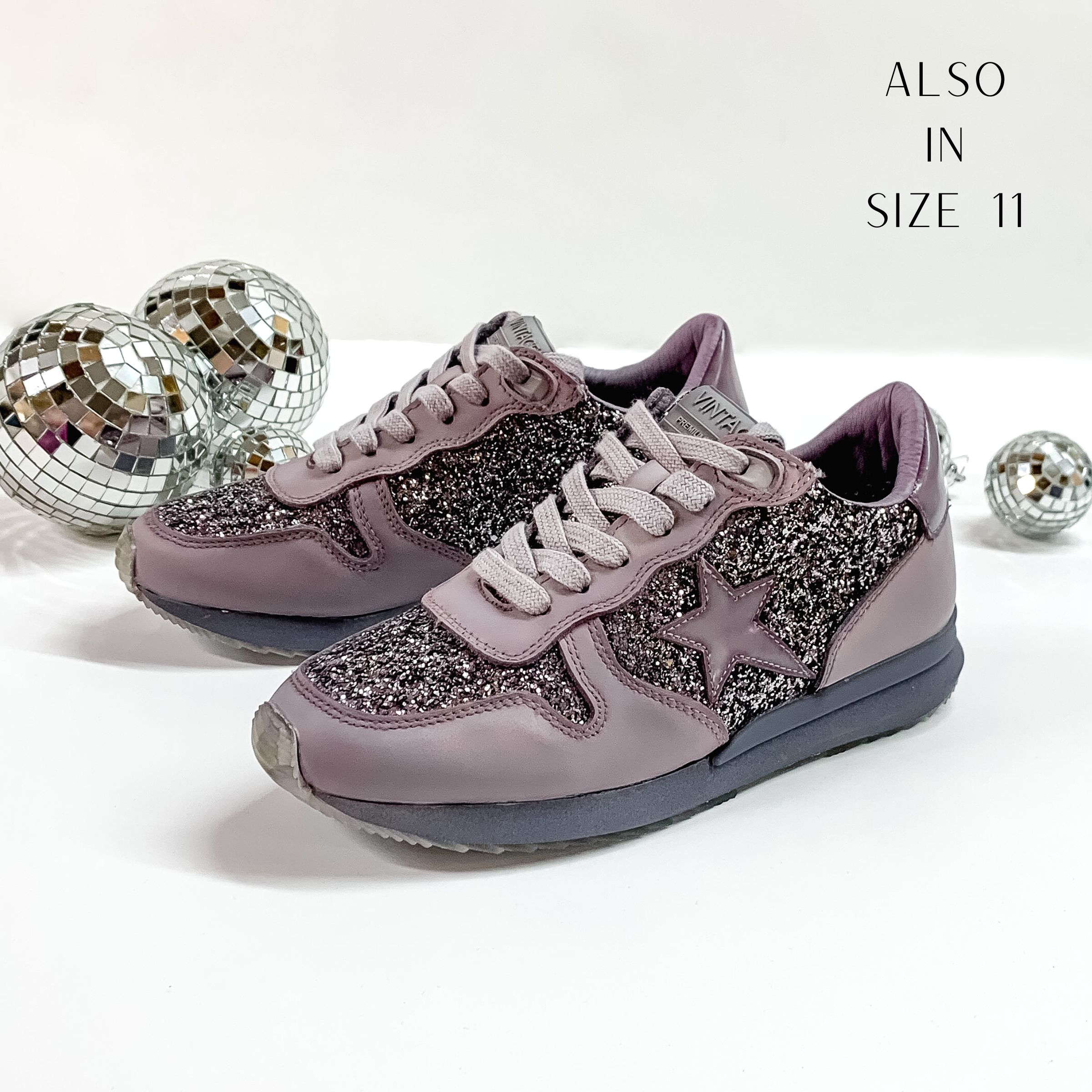Grey purple colored tennis shoes and shoelaces. The tennis shoes also have a grey purple star patch on the side of the shoe and grey purple glitter throughout. These shoes are pictured on a white background with disco balls behind them. 