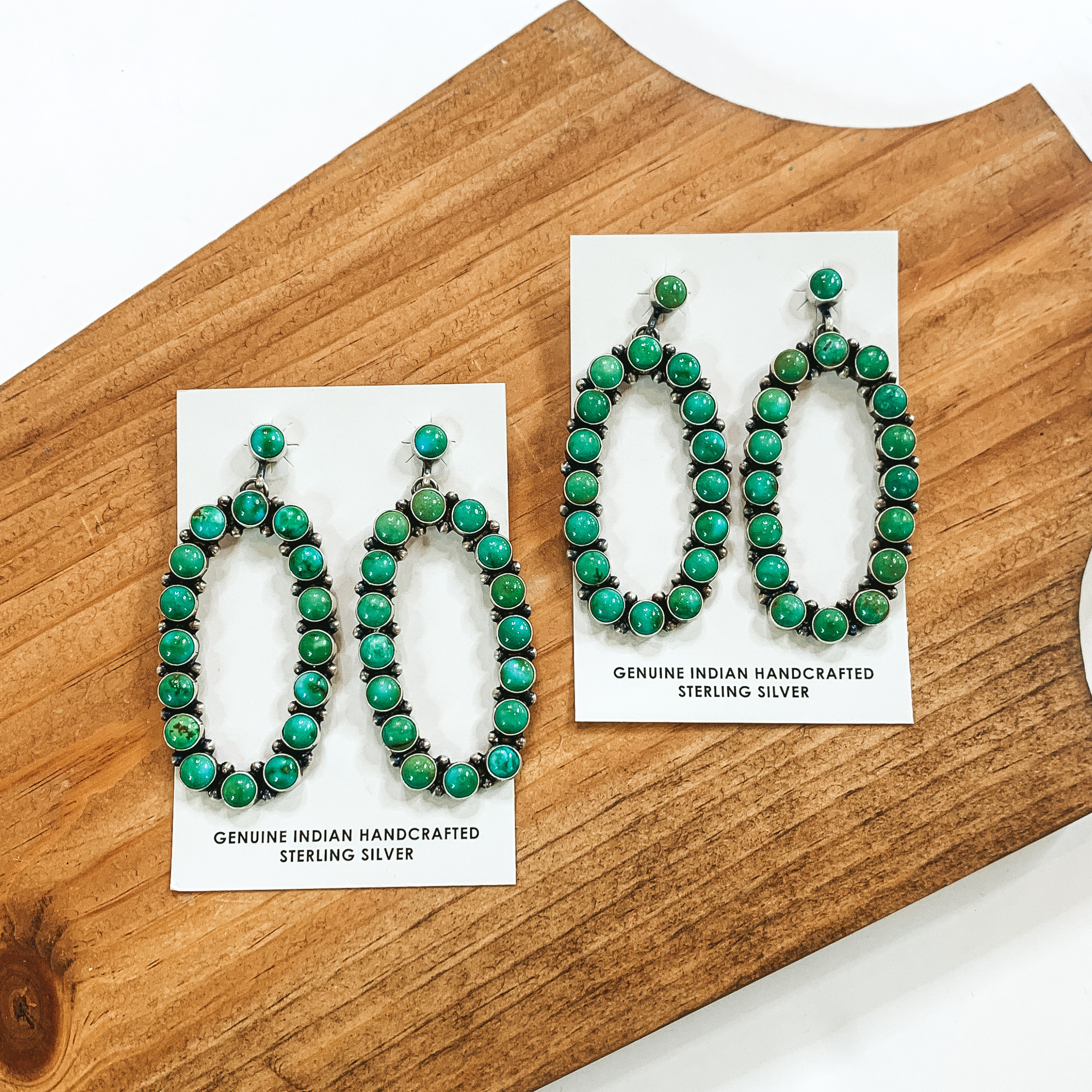 Open oval drop earrings that have green colored stones. There are two pairs of these earrings pictured on a wood block on a white background. 