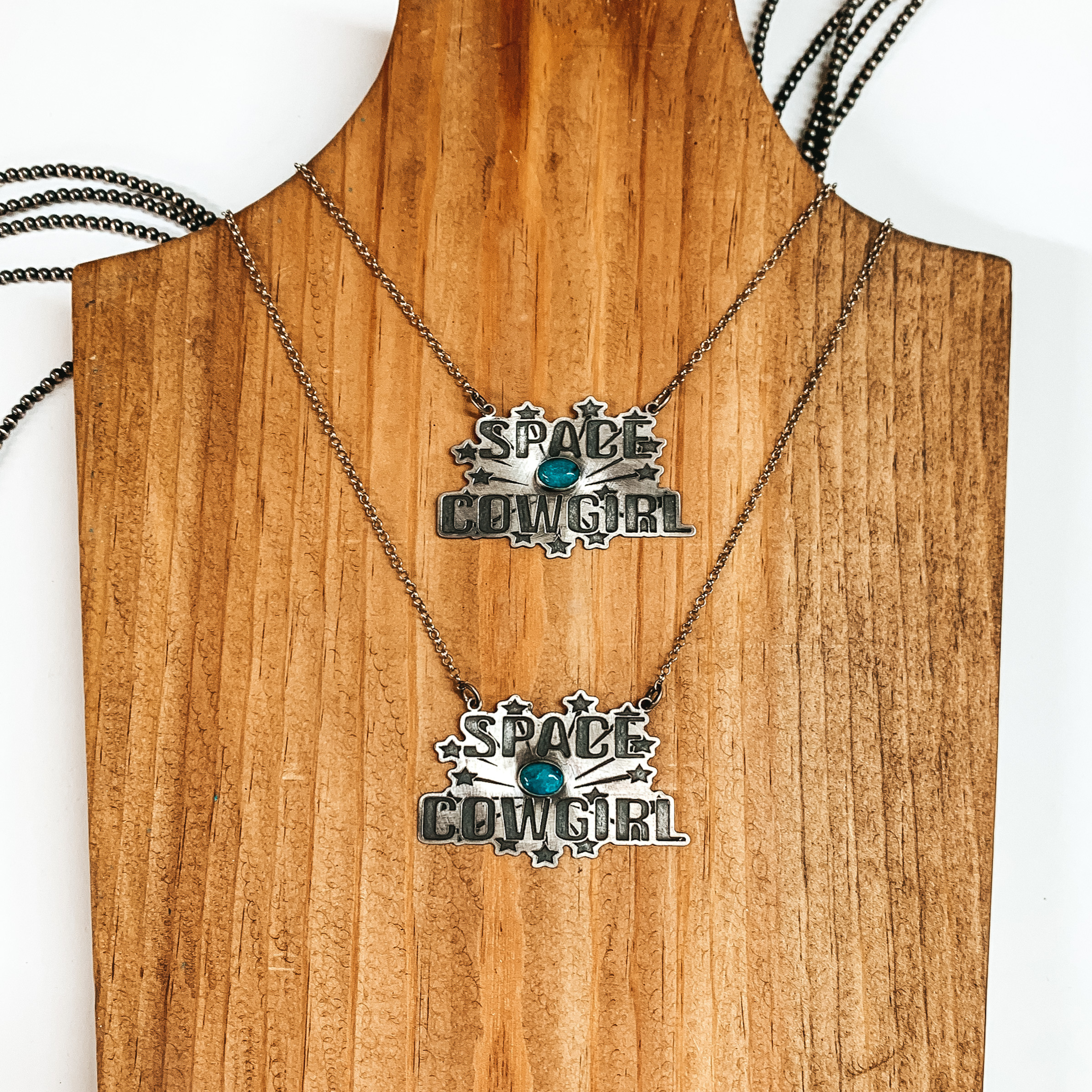 Silver chain necklace with a silver pendant and small turquoise stone. The silver pendant has the words "SPACE COWGIRL" engrave on it with stars surrounding it. This necklace is pictured on a wood block on a white background. 