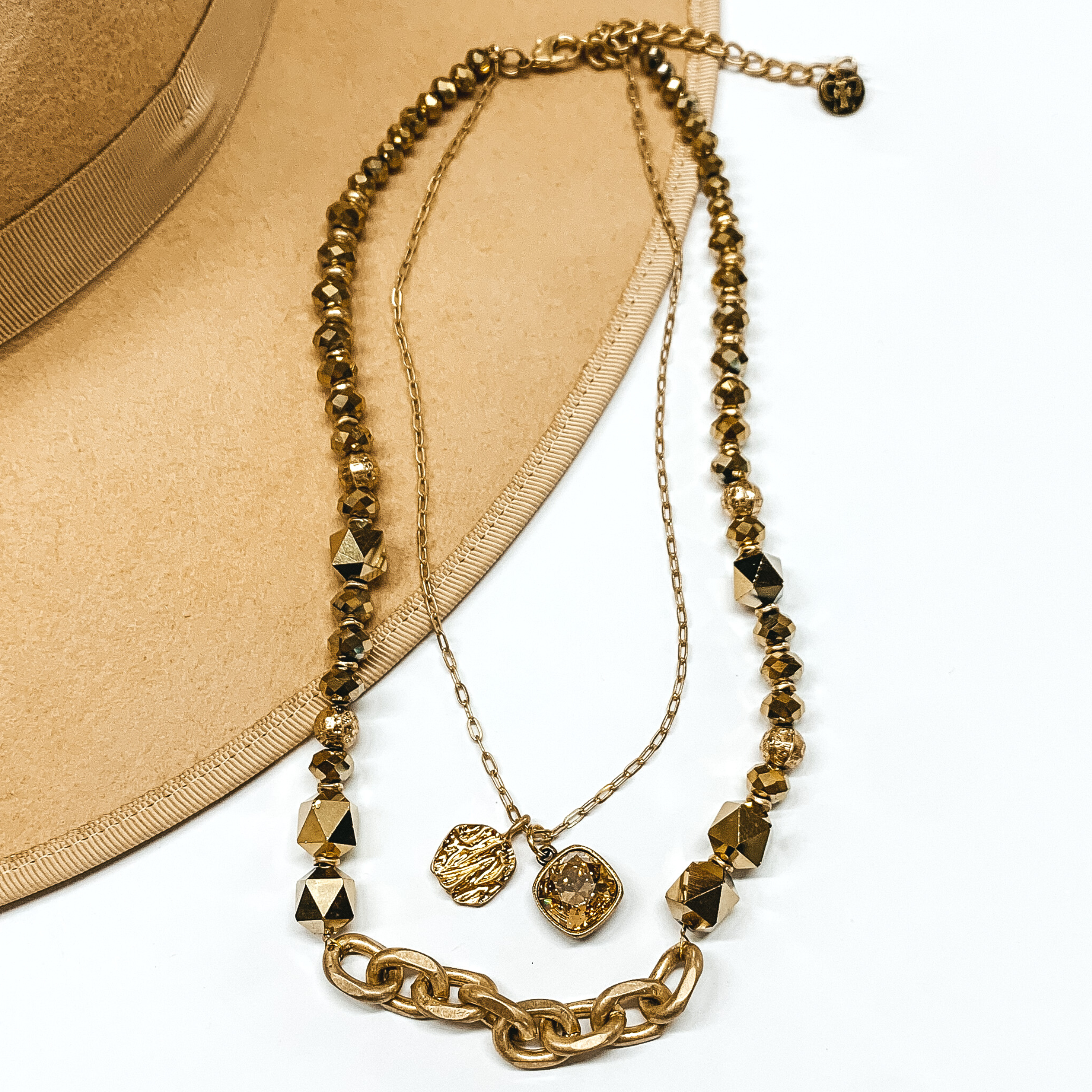 This is a two strand necklace that has one short dainty chain and a longer gold crystal beaded strand with a gold chain segment. The shorter chain includes a gold coin charm and golden shadow cushion cut crytal charm. This necklace is pictured partially laying on a tan brim on a white background. 