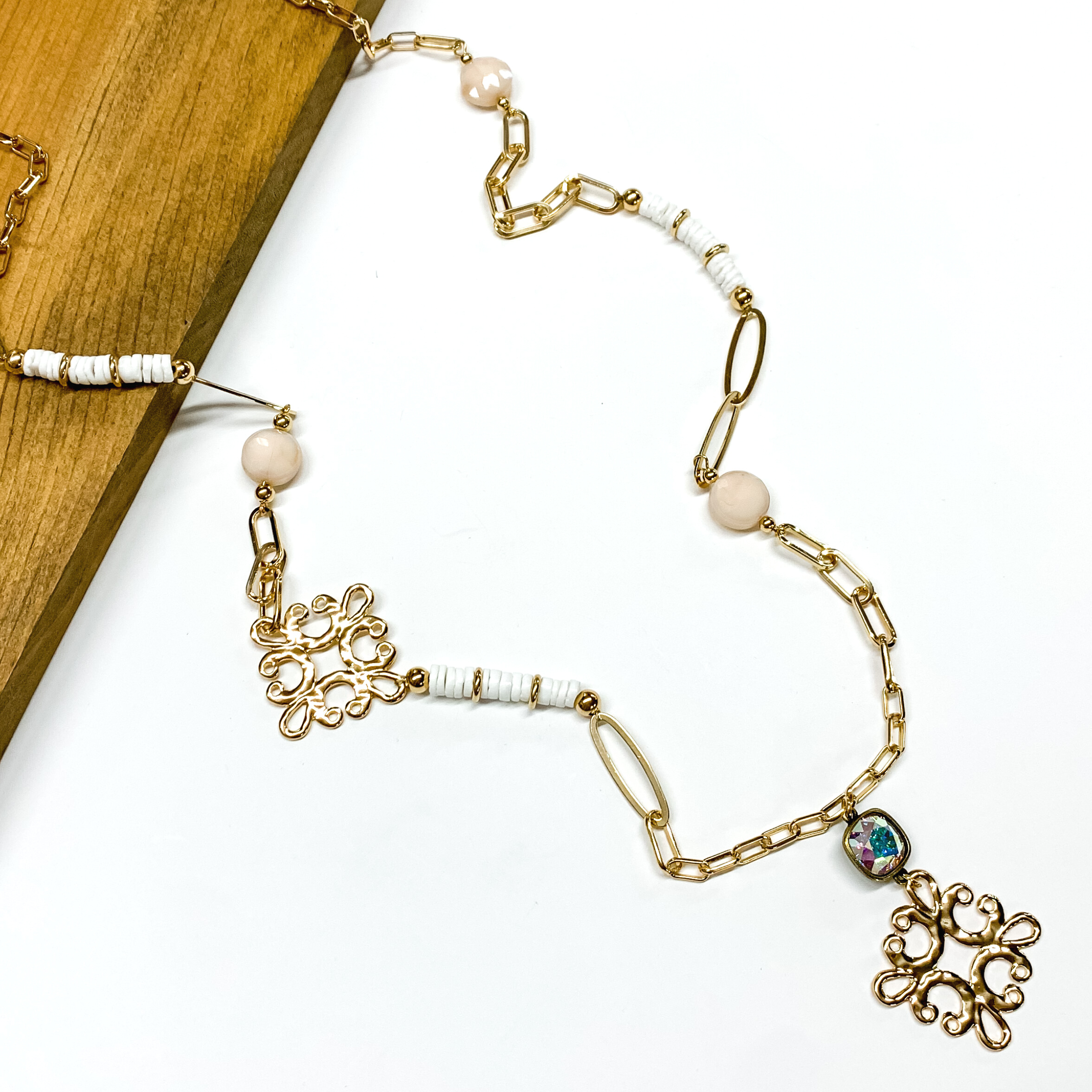 Gold paperclip chain and white beaded necklace. This necklace also includes an ab crystal and gold quatrefoil drop. This necklace is pictured partially laying on a brown block on a white background. 