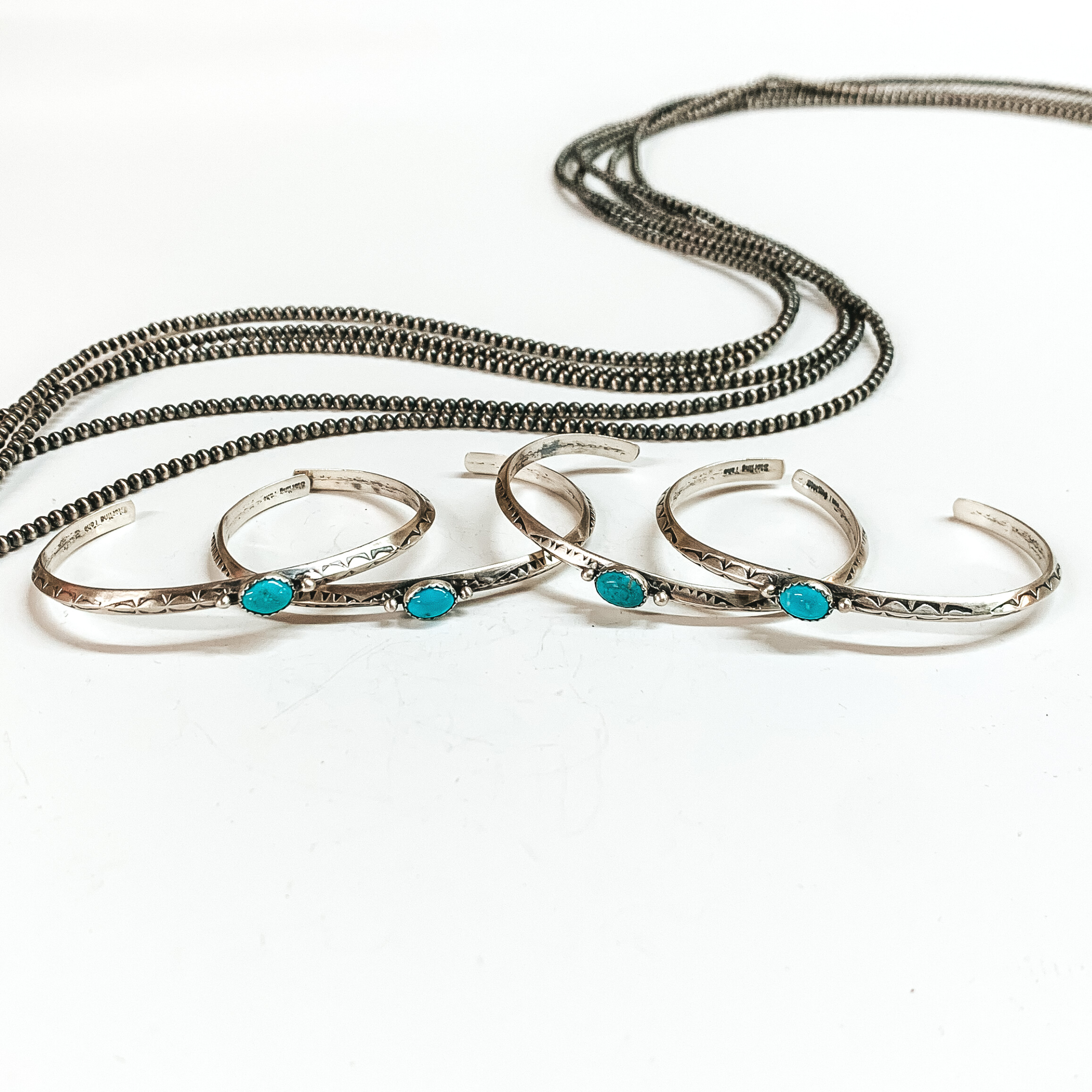 Four silver cuff bracelets that have detailed engraving and center turquoise stone. These bracelets are pictured laying on top of each other on a white background with silver beads behind them. 