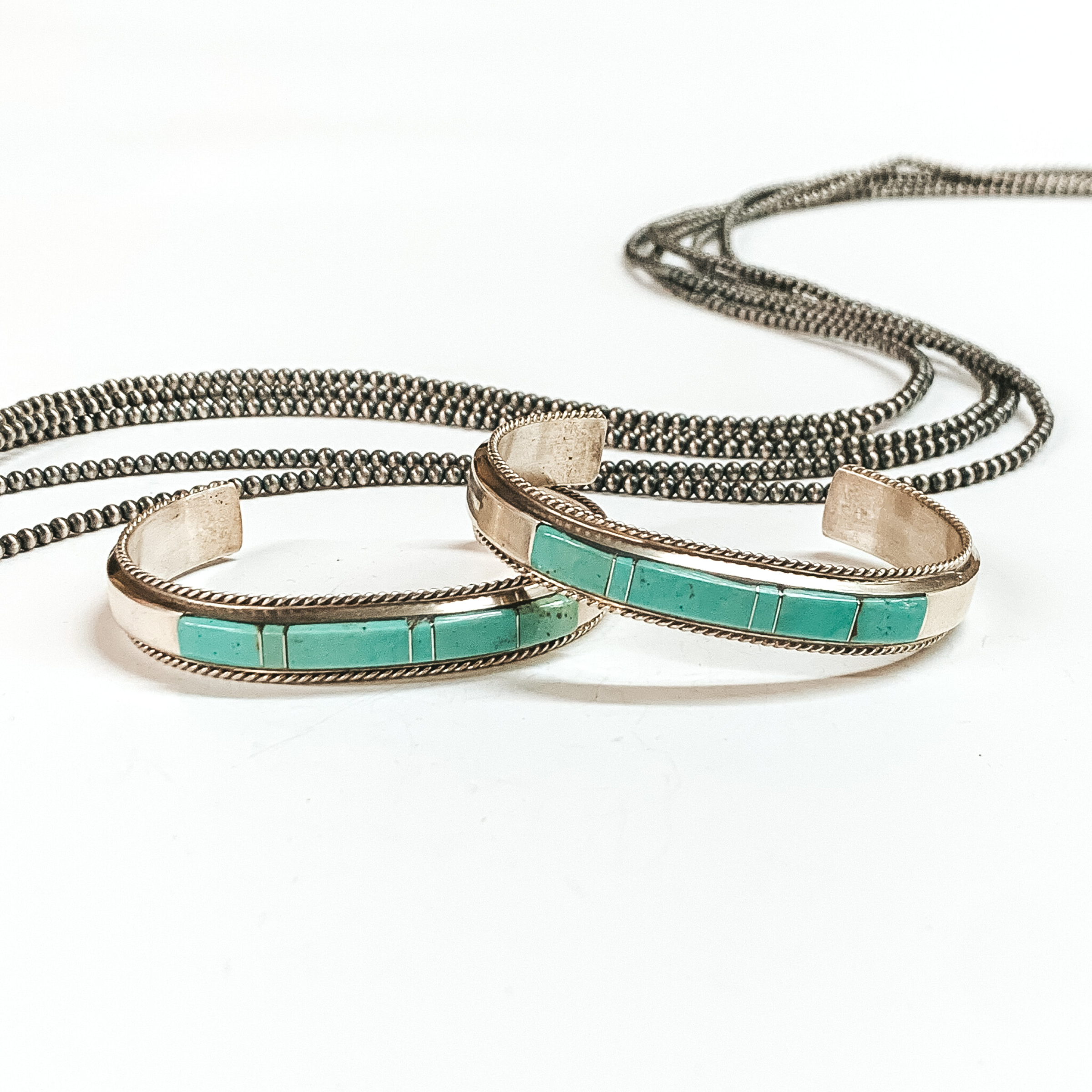Two sterling silver cuff bracelets with six turquoise stone inlay. These bracelets are pictured on a white background with silver beads above the bracelets. 