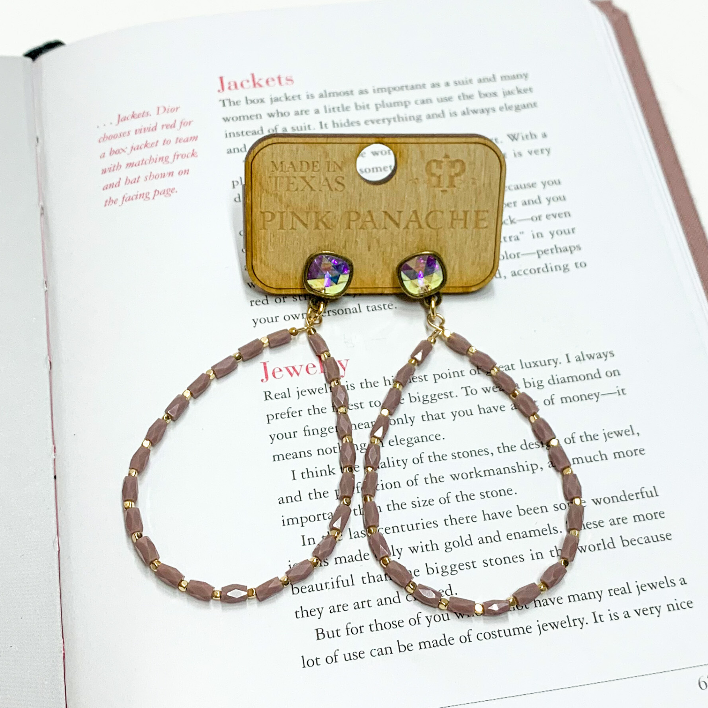 Ab cushion cut crystals with a hanging teardrop. The teardrop has lavender and gold beads. This pair of earrings are pictured on an open book. 