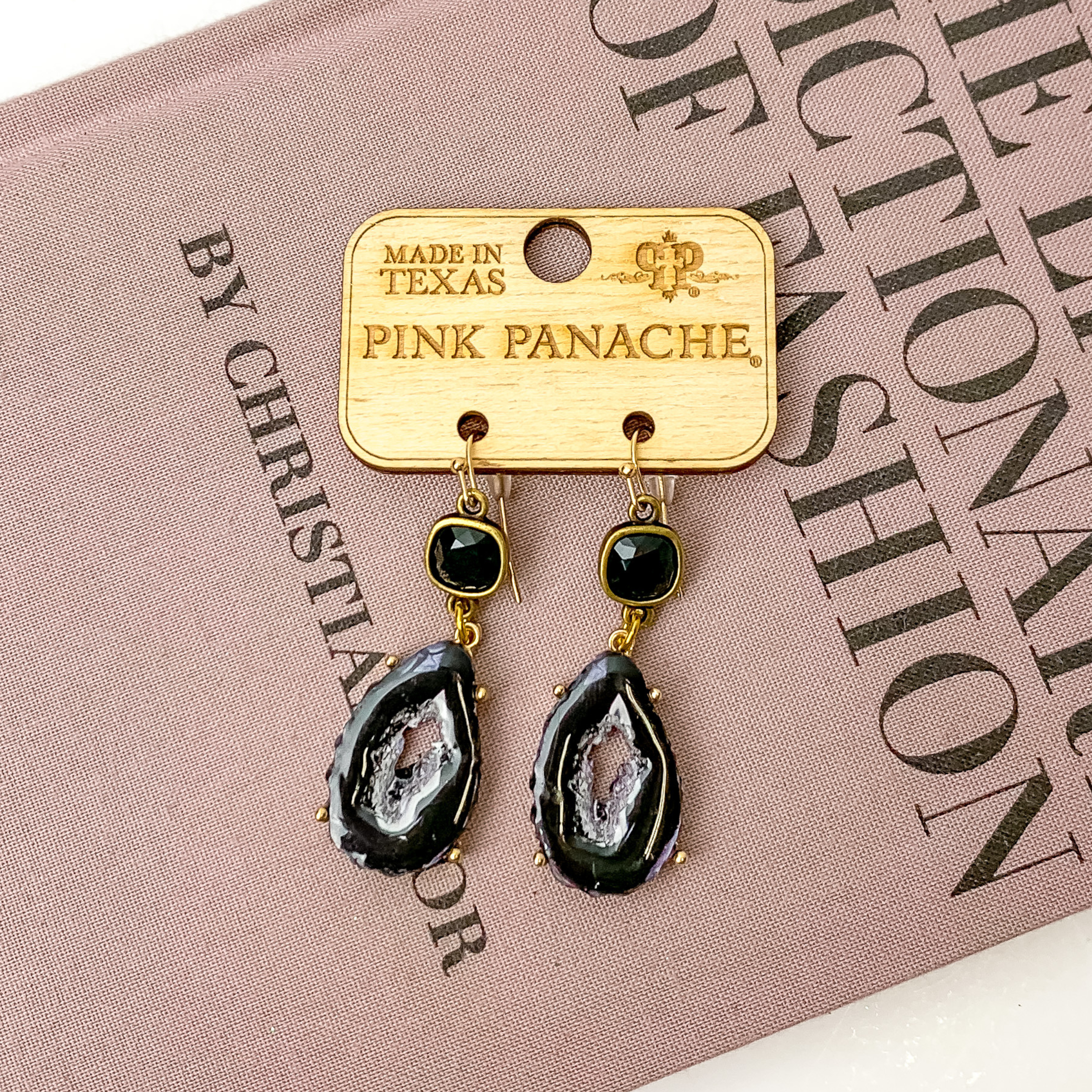 Black cushion cut crystal earrings with a hanging black, druzy stone pendant. These earrings are pictured on a mauve book on a white background. 
