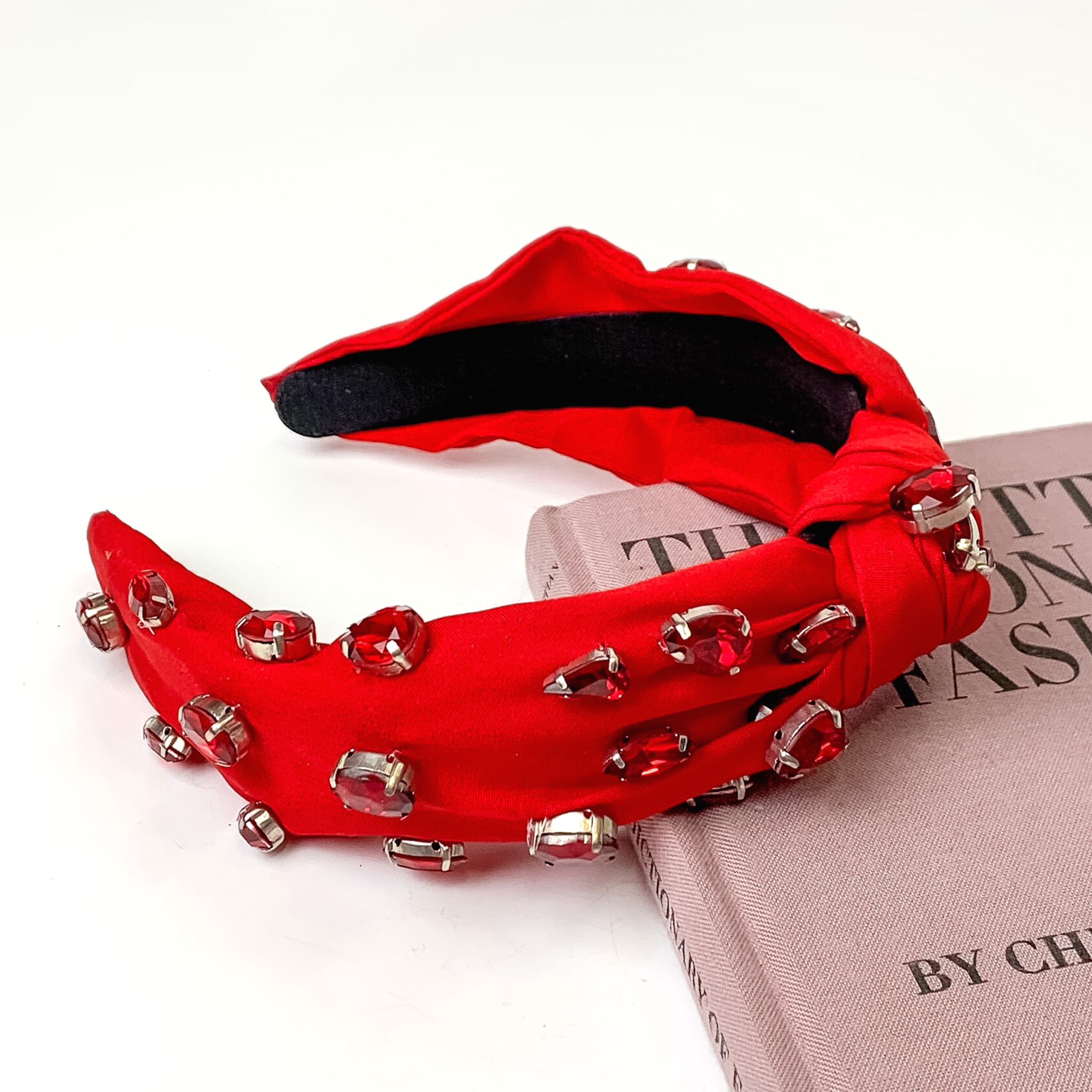 Red colored knot headband with different shaped red crystals. This headband is pictured partially laying on a mauve book on a white background.