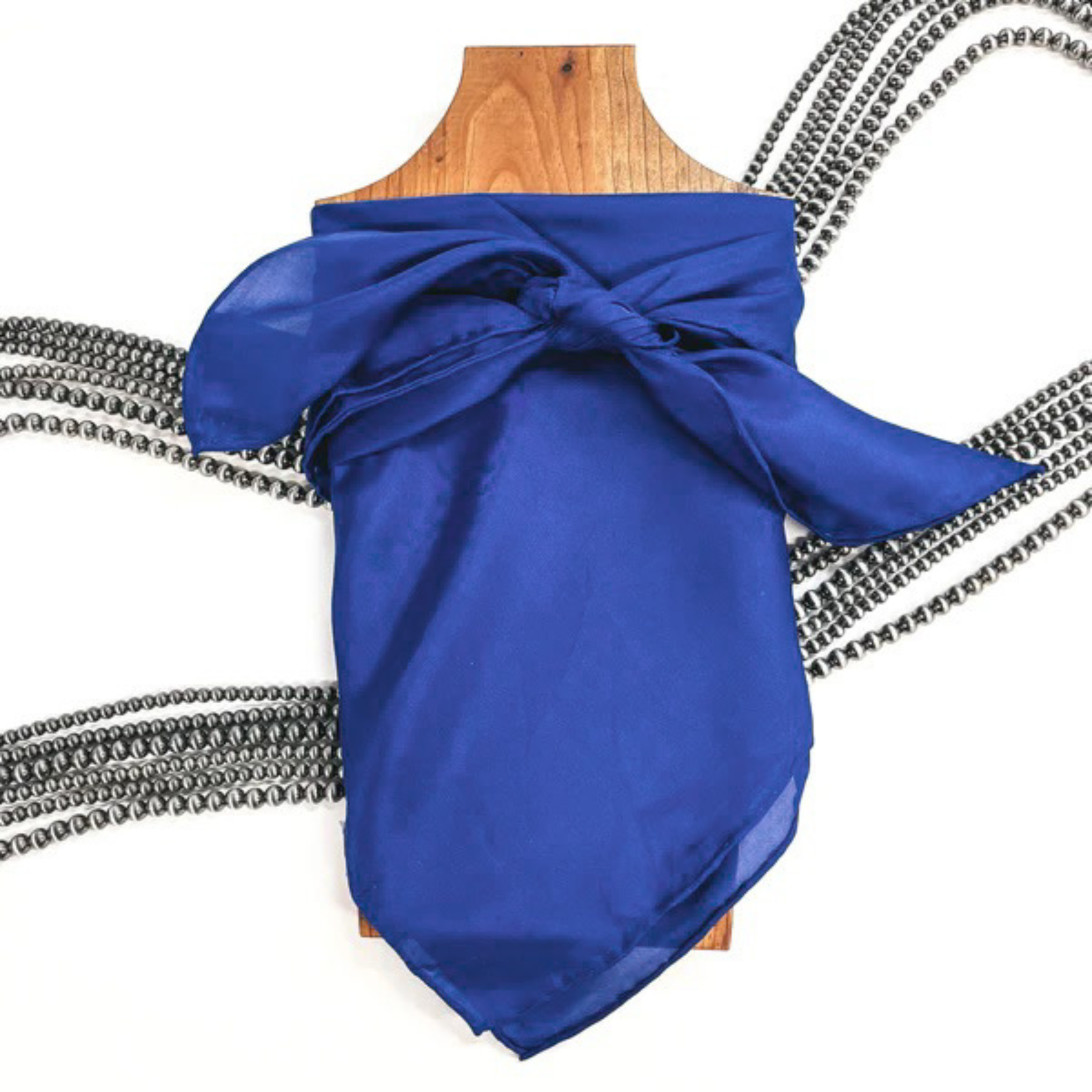 This is a royal blue silk wild rag, this wild rag is placed on a brown necklace board. This wild rag is taken on a white background and with silver Navajo pearls in the back as decor.