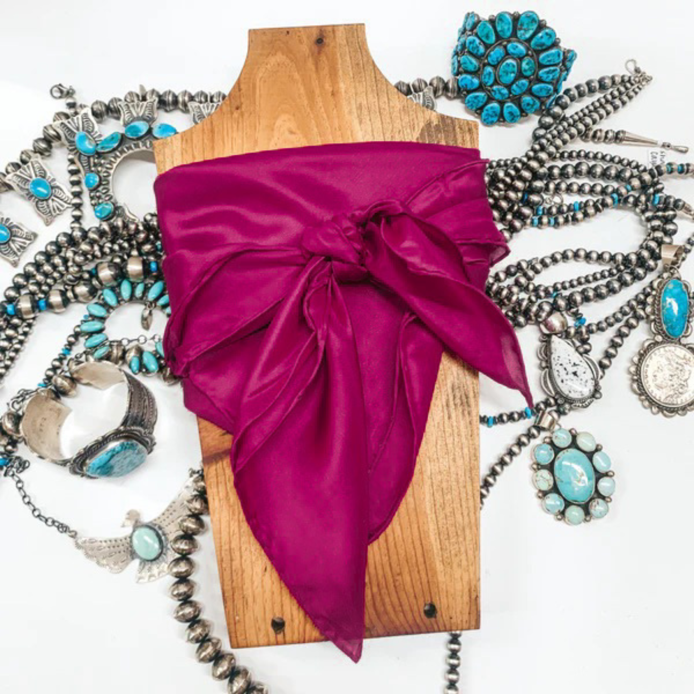 This is a fuchsia silk wild rag, this wild rag is pictured wrapped around a brown necklace board. This wild rag is pictured on a white background and with silver and turquoise Navajo jewelry behind it as decoration.