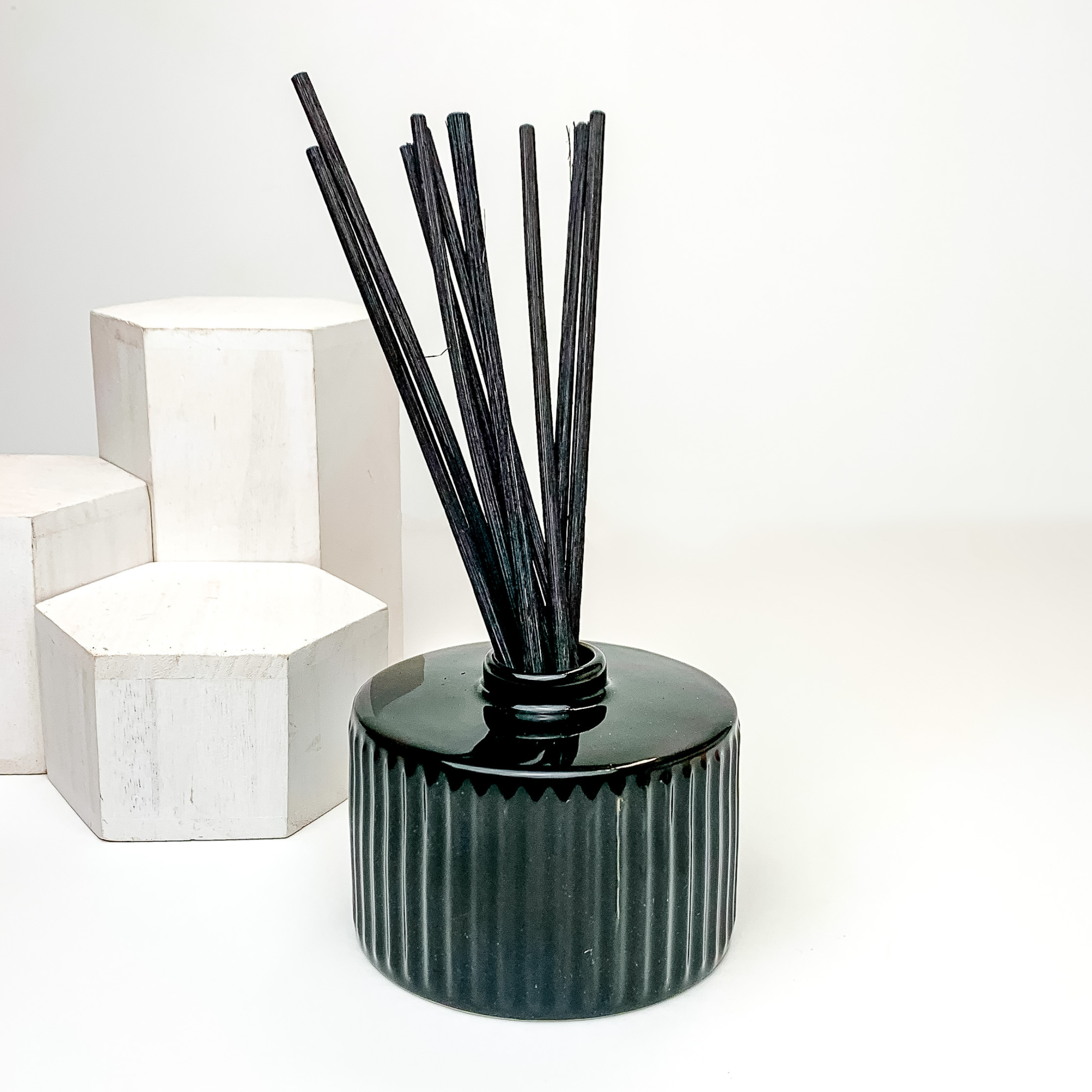 Black diffuser holder with black reeds sticking out of the top. This diffuser is pictured on a white background with white blocks on the left side of the candle. 