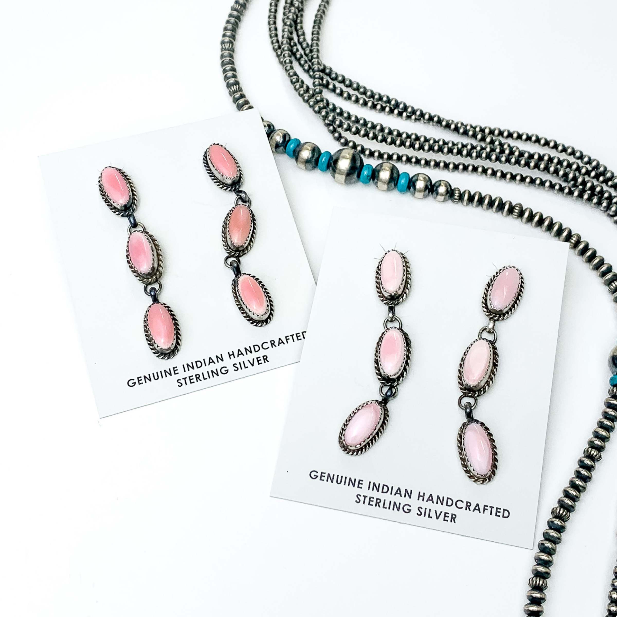 Pictured are two pairs of silver and oval drop earrings. These earrings include three pink oval stones with silver outlines. These earrings are pictured on a white background with silver beads on the right side of the picture. 