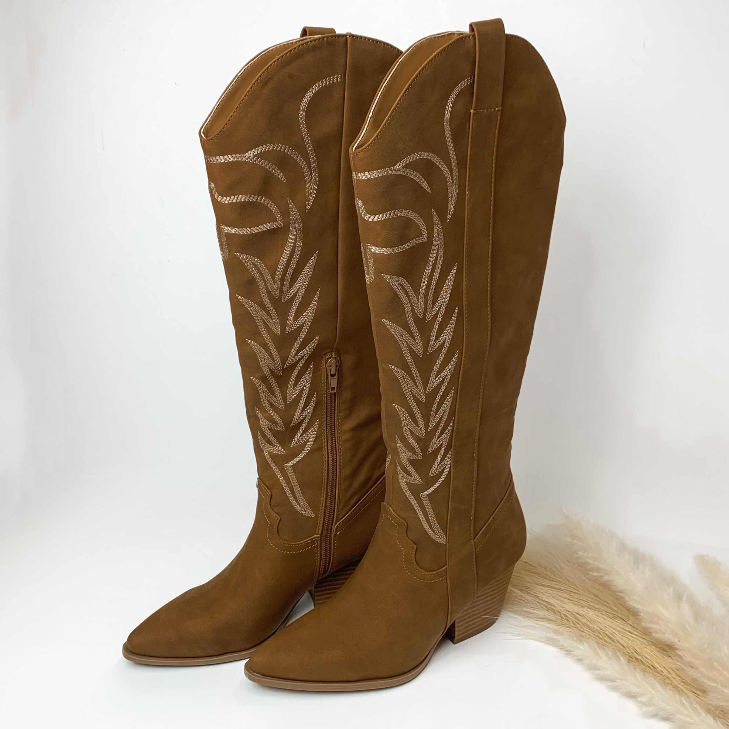 Camel brown tall boots with ivory western stitching and tan heel. These boots are pictured on a white background with some ivory pompous grass on the right side of the boots. 