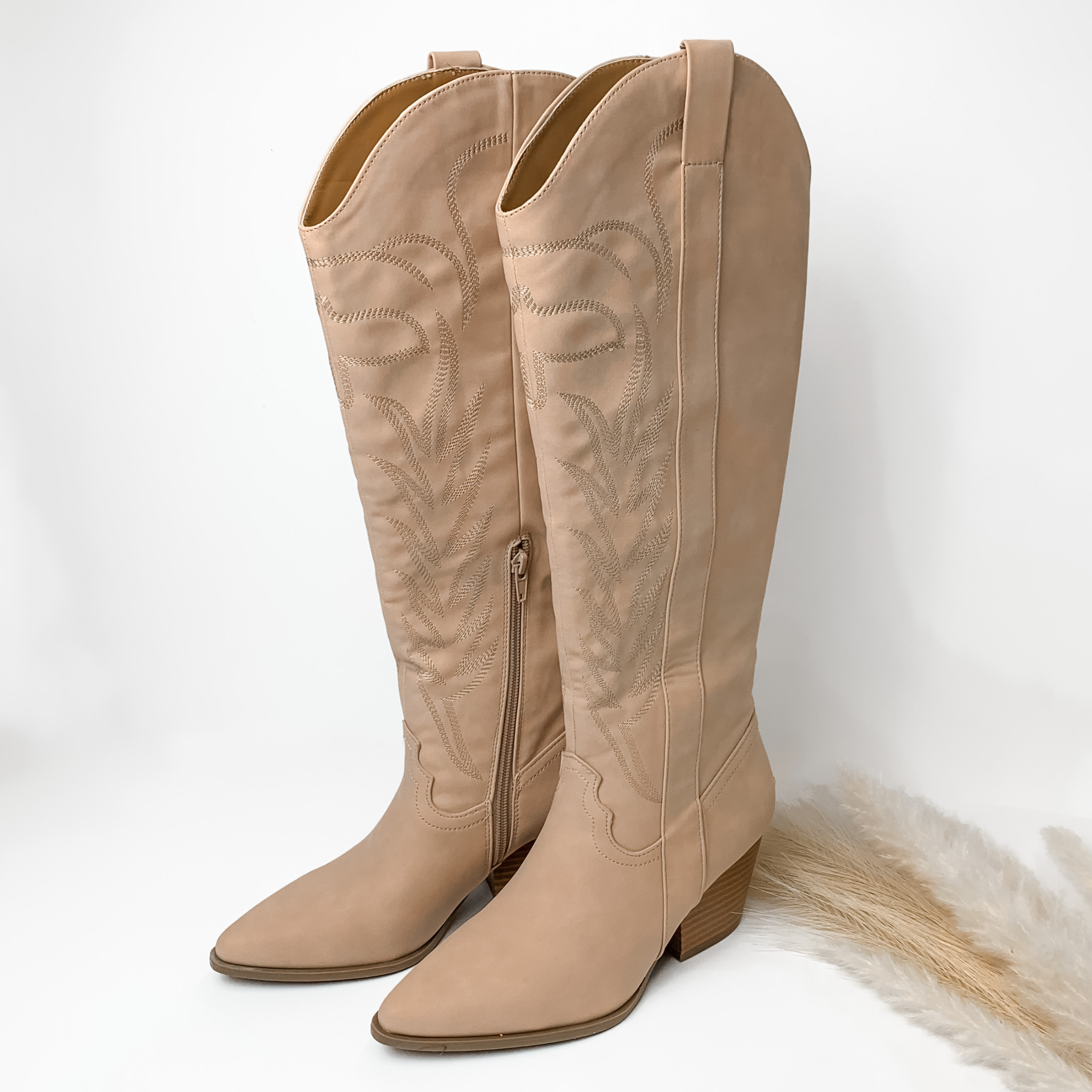 Nude tall boots with nude western stitching and tan heel. These boots are pictured on a white background with some ivory pompous grass on the right side of the boots. 