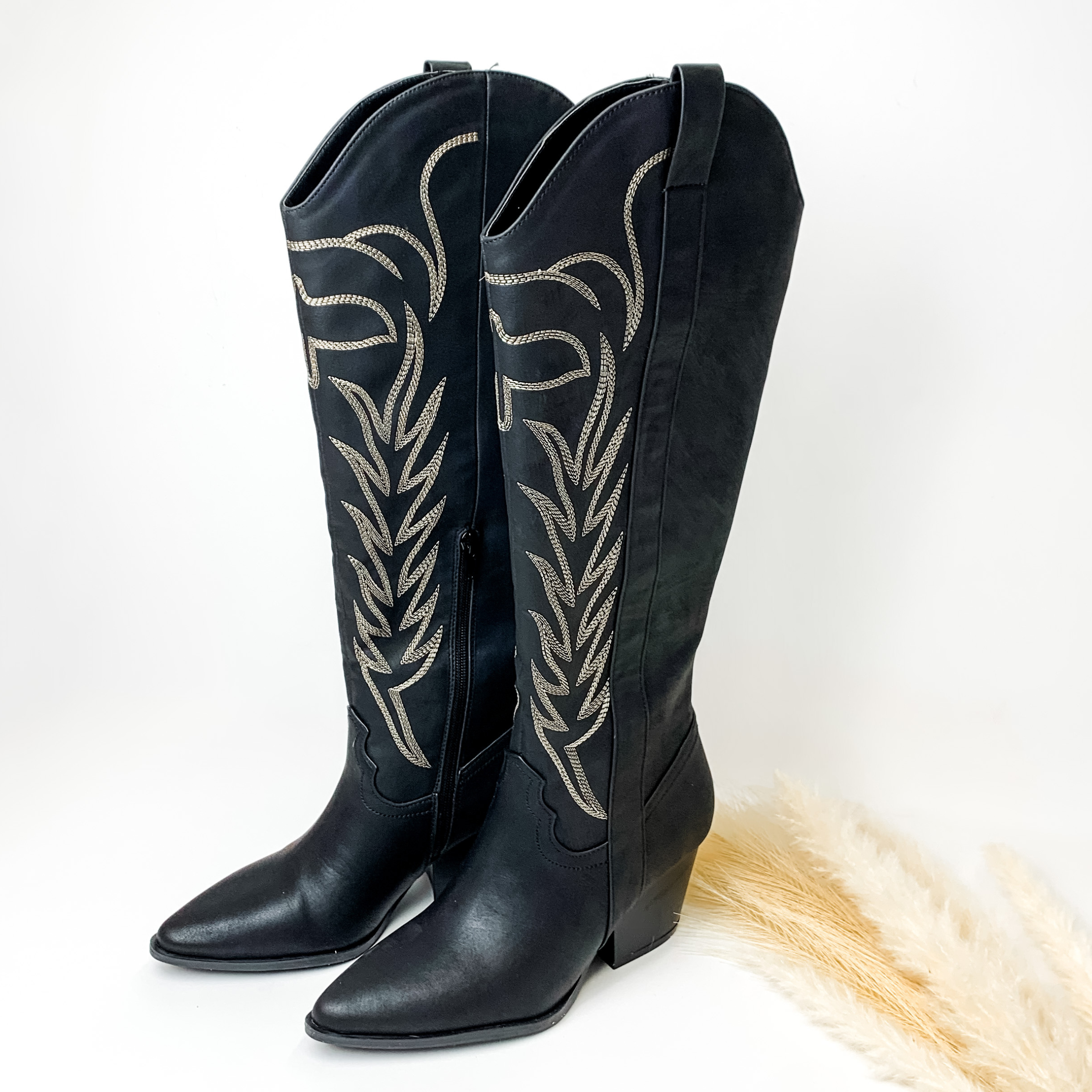 Black tall boots with ivory western stitching and tan heel. These boots are pictured on a white background with some ivory pompous grass on the right side of the boots. 