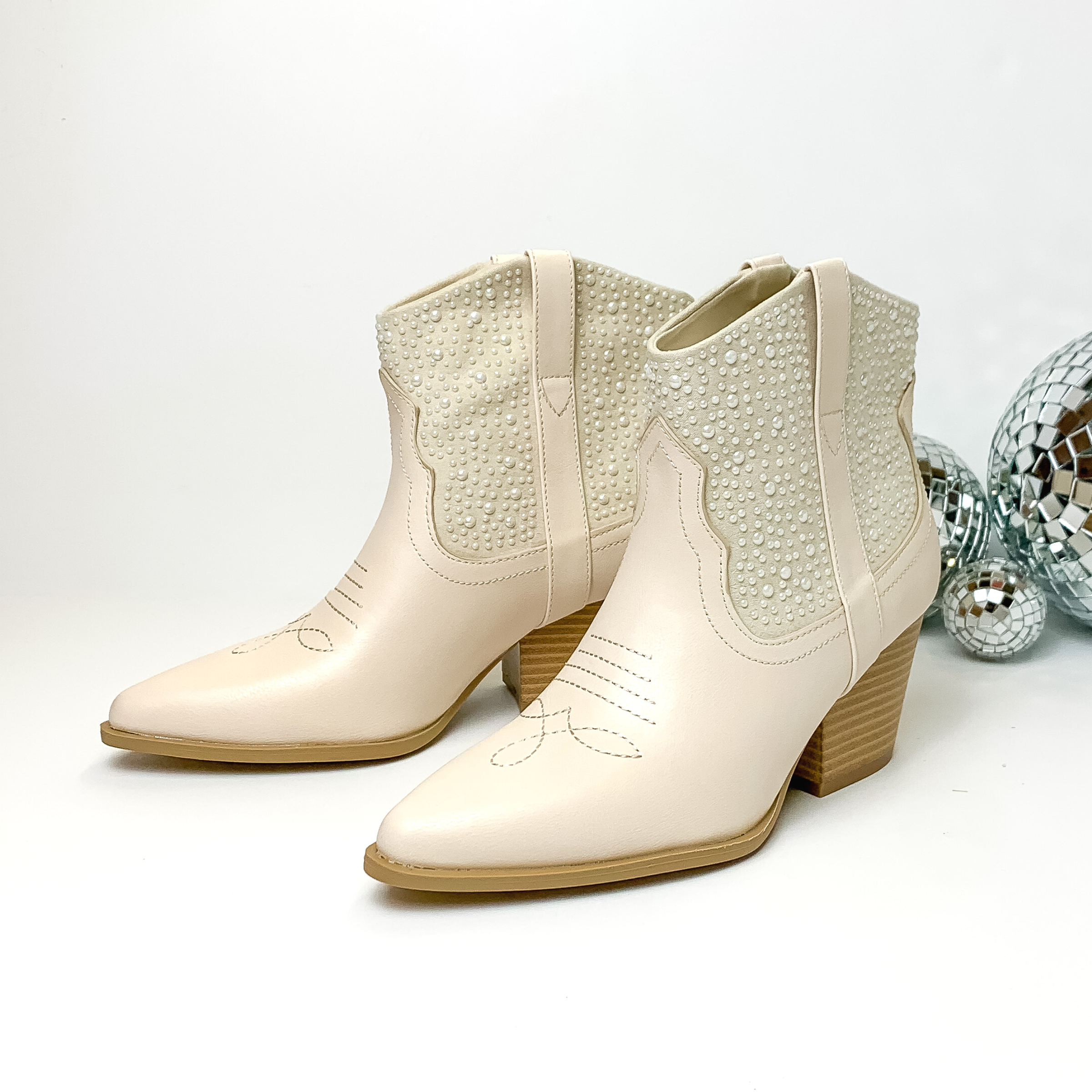 Ivory ankle boots with white pearl embellishment and tan heel. These boots are pictured on a white background with disco balls to the right of the boots.