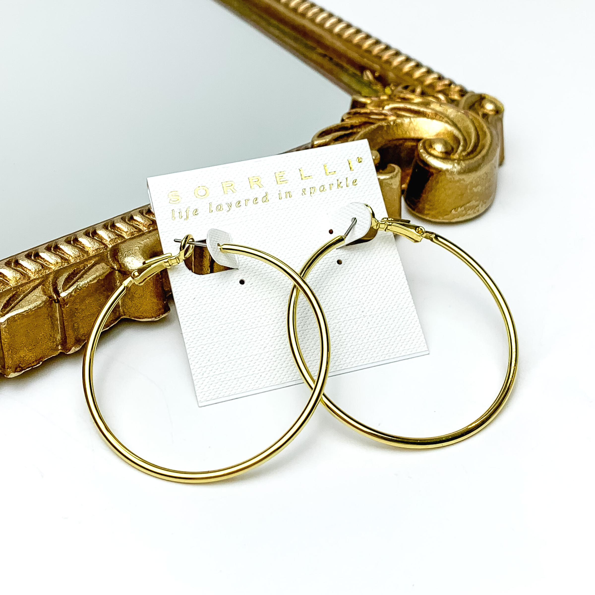 Pair of gold hoop earrings pictured on a white earrings holder. These earrings are pictured in front of a gold mirror on a white background. 