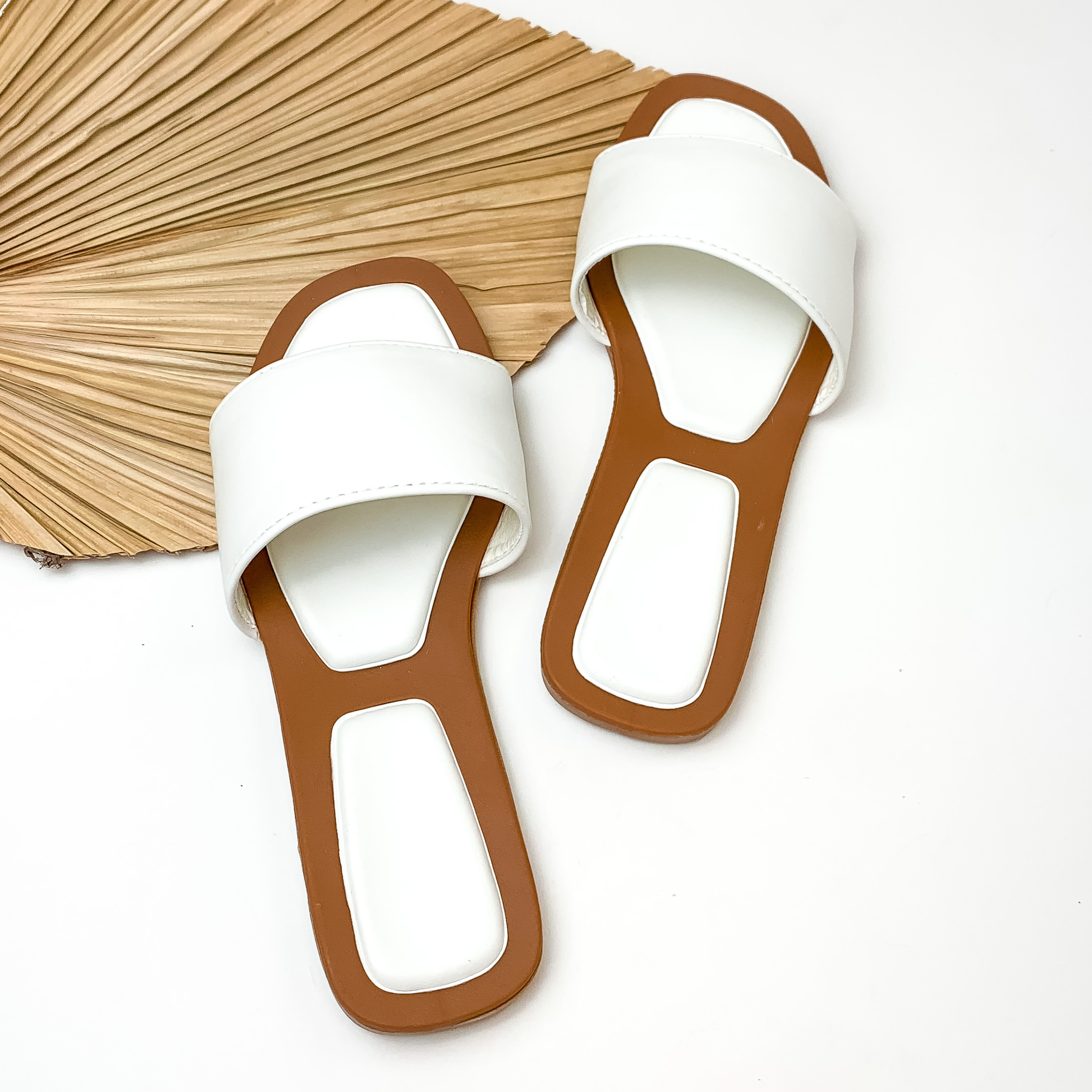 Pictured are white, single wide strap sandals. These sandals have a tan sole with white comfort spots. these sandals are pictured on a white background with tan palm leaf.