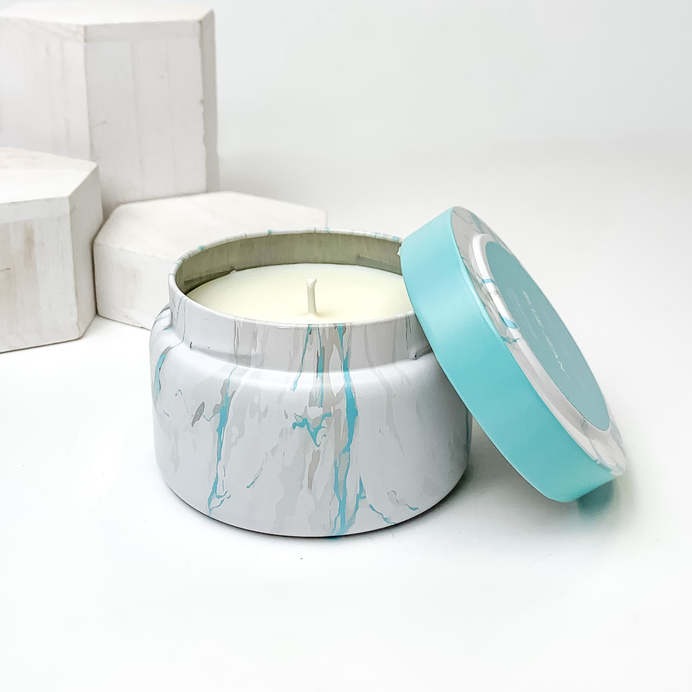 White, grey and turquoise marble candle with the lid leaning on the side of the candle. This candle is pictured in front of white blocks on a white background. 