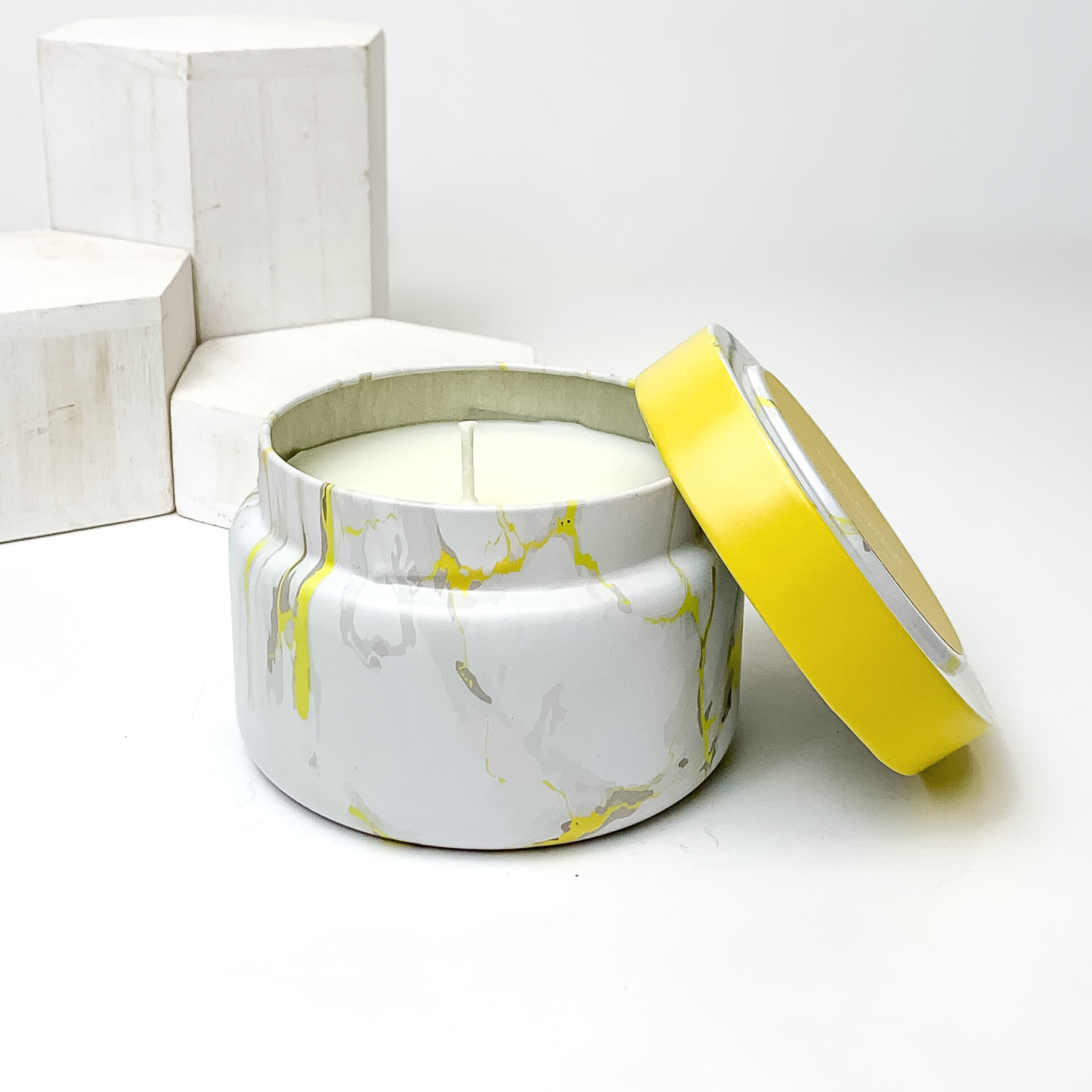 White, grey and yellow marble candle with the lid leaning on the side of the candle. This candle is pictured in front of white blocks on a white background. 