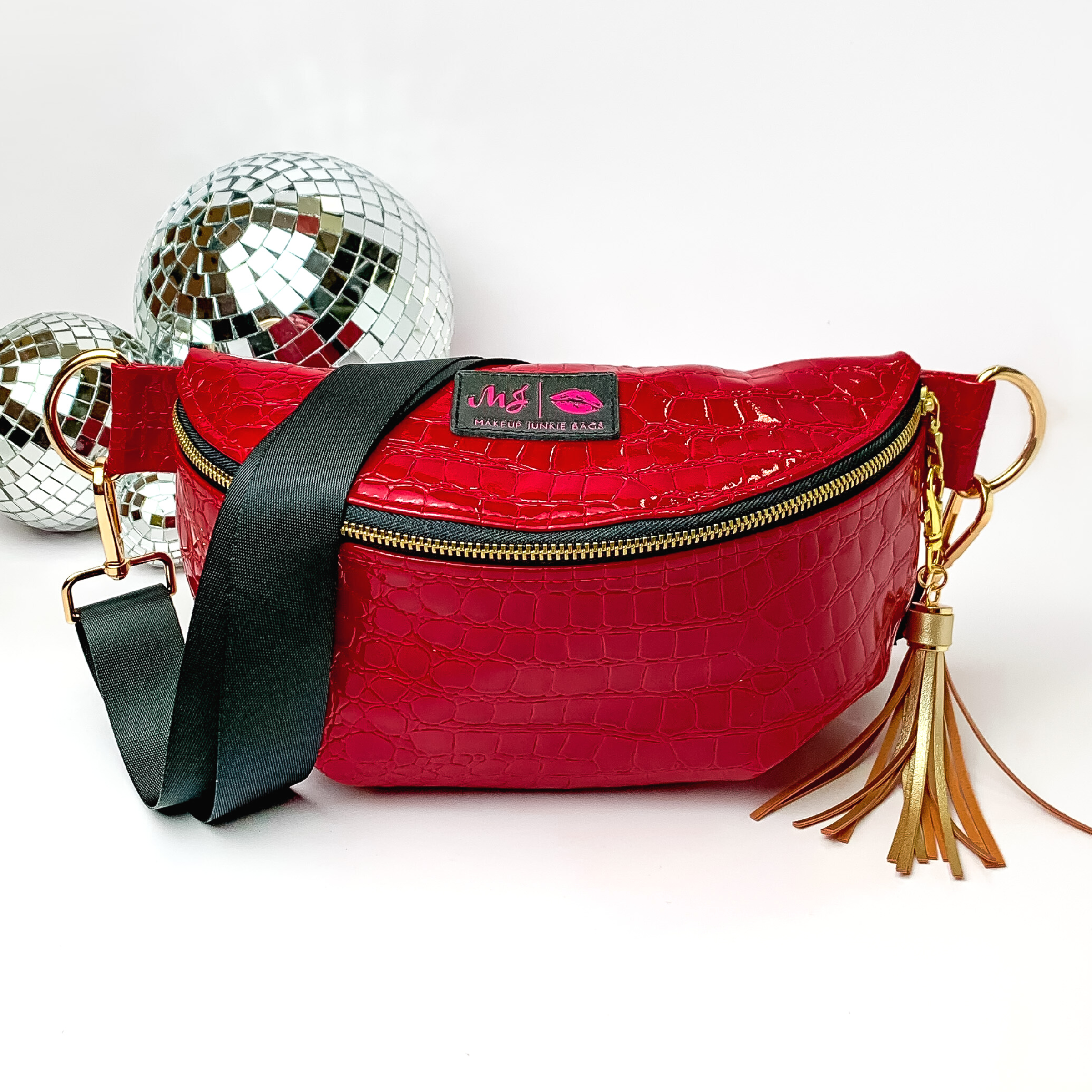Cherry red, croc print fanny pack with a black strap. This bag includes gold accents, a gold zipper, and a gold tassel. This bag also has a pink stitched logo for Makeup Junkie. This fanny pack is pictured on a white background in front of disco balls. 