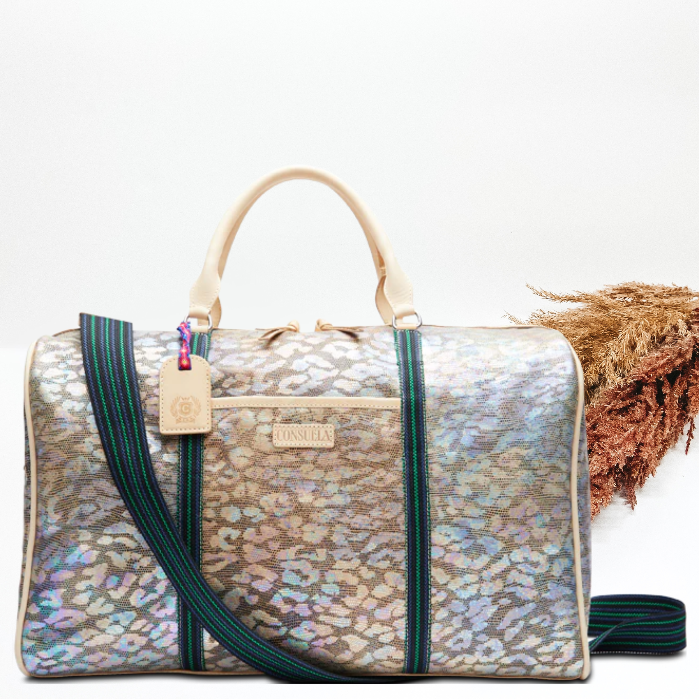 Pictured is a duffle bag with iridescent, multicolor leopard print design. This duffle bag includes short, light tan handles and a long, striped strap. This bag is pictured on a white background with pompous grass on the left side of the picture. 