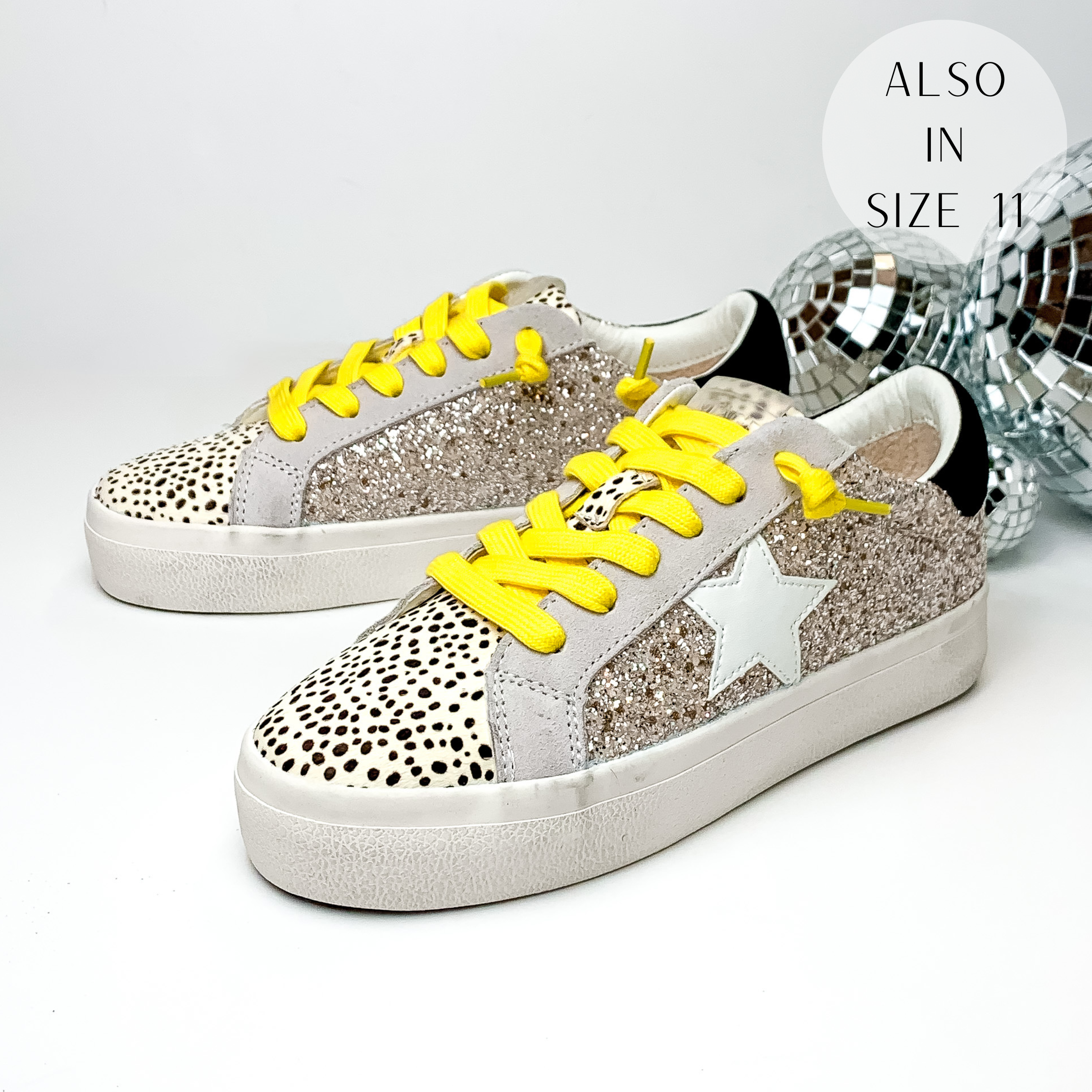 Pictured are tennis shoes with silver glitter on the sides and a cheetah print on the tongue down to the toe. These shoes also include a black patch on the heel, a white star emblem on the side of the shoe, and yellow shoe laces. These shoes are pictured on a white background with disco balls behind them on the right hand side.