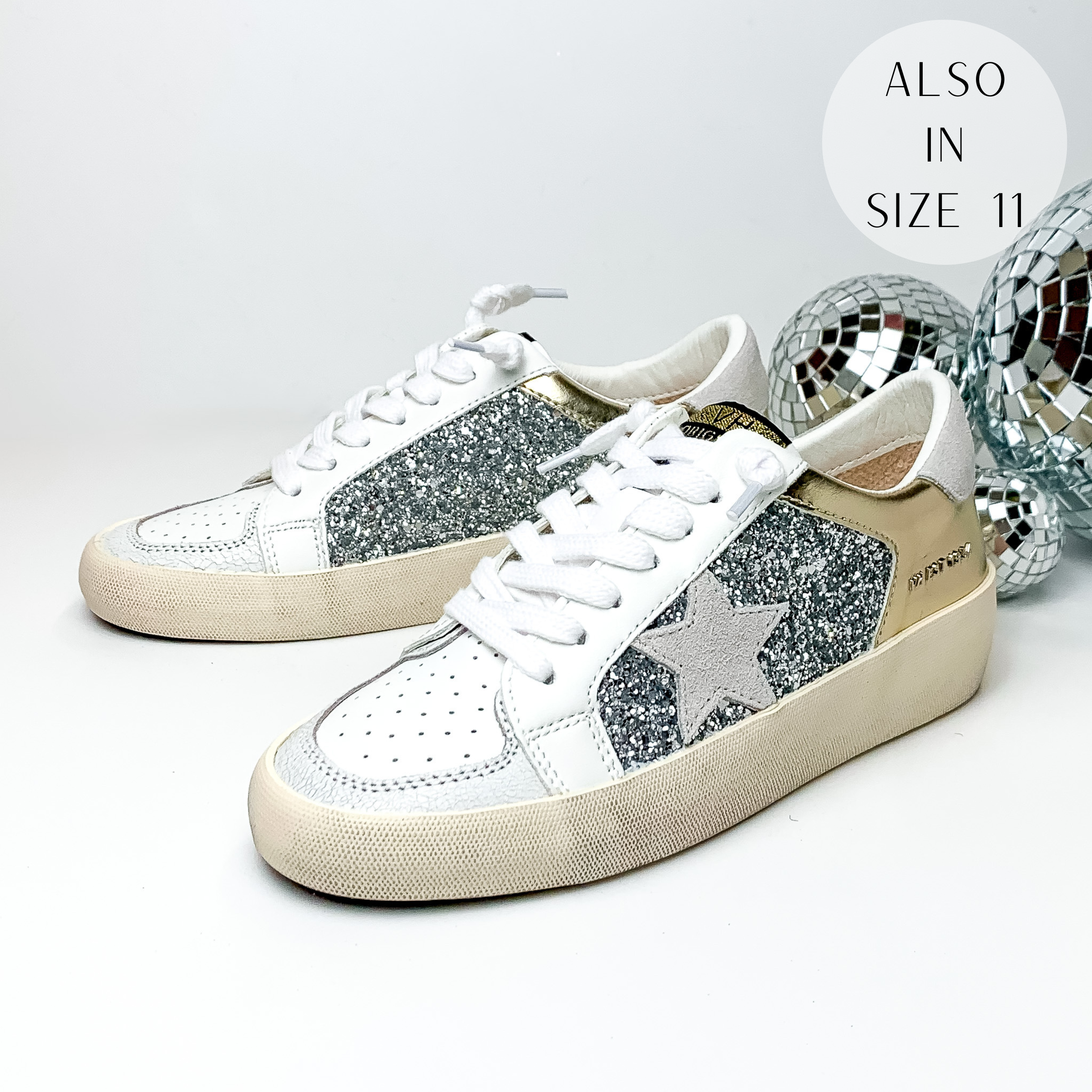 Pictured are white tennis shoes with silver glitter on the sides and a mettalic gold patch on the side and back of the shoe. These shoes also include a light grey patch on the heel, a light grey star emblem on the side of the shoe. These shoes are pictured on a white background with disco balls behind them on the right hand side.