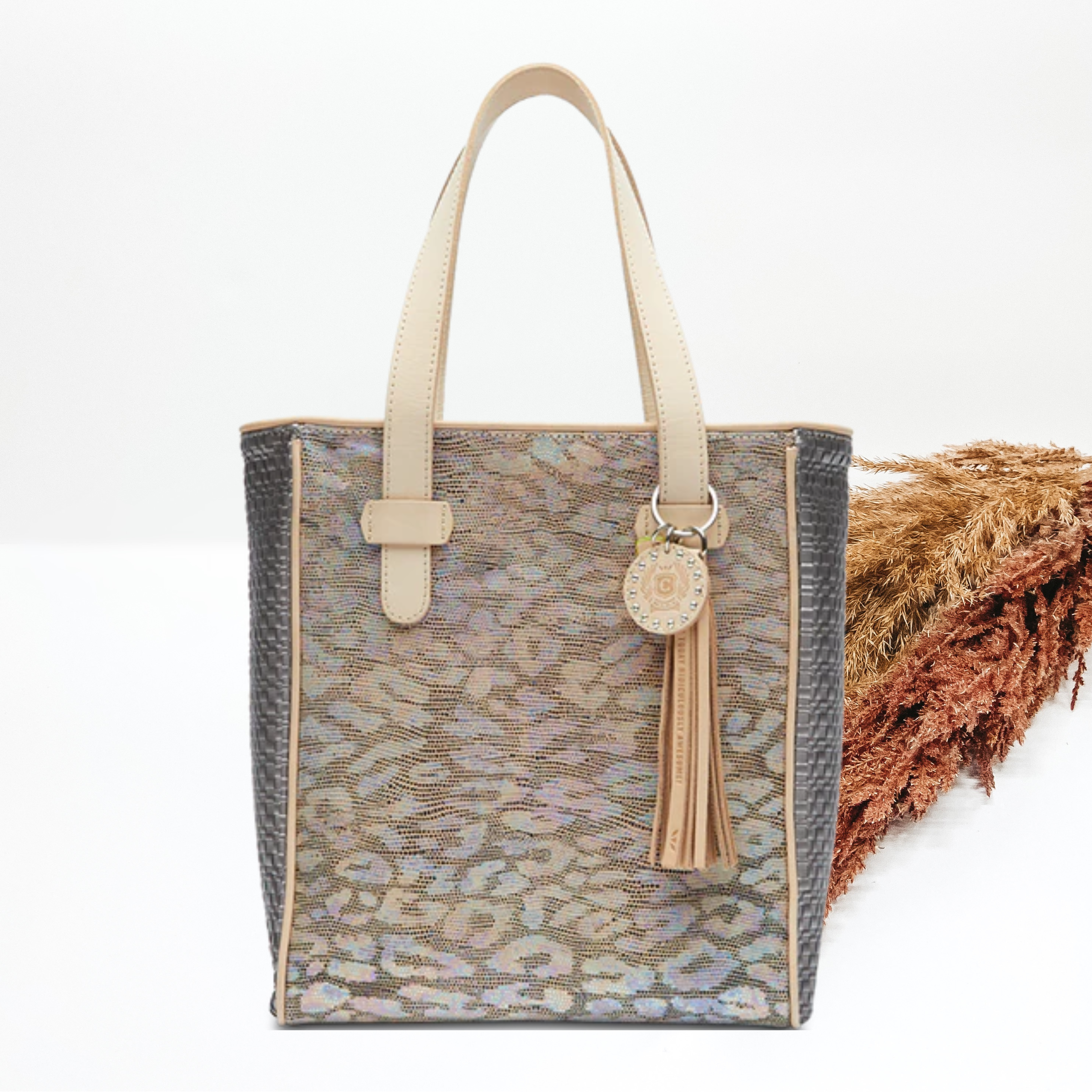 Iridescent leopard print tote bag in multicolor. This tote has light tan handles, silver sides, and a light tn tassel charm. this tote is pictured in front of pompous grass on a white background. 