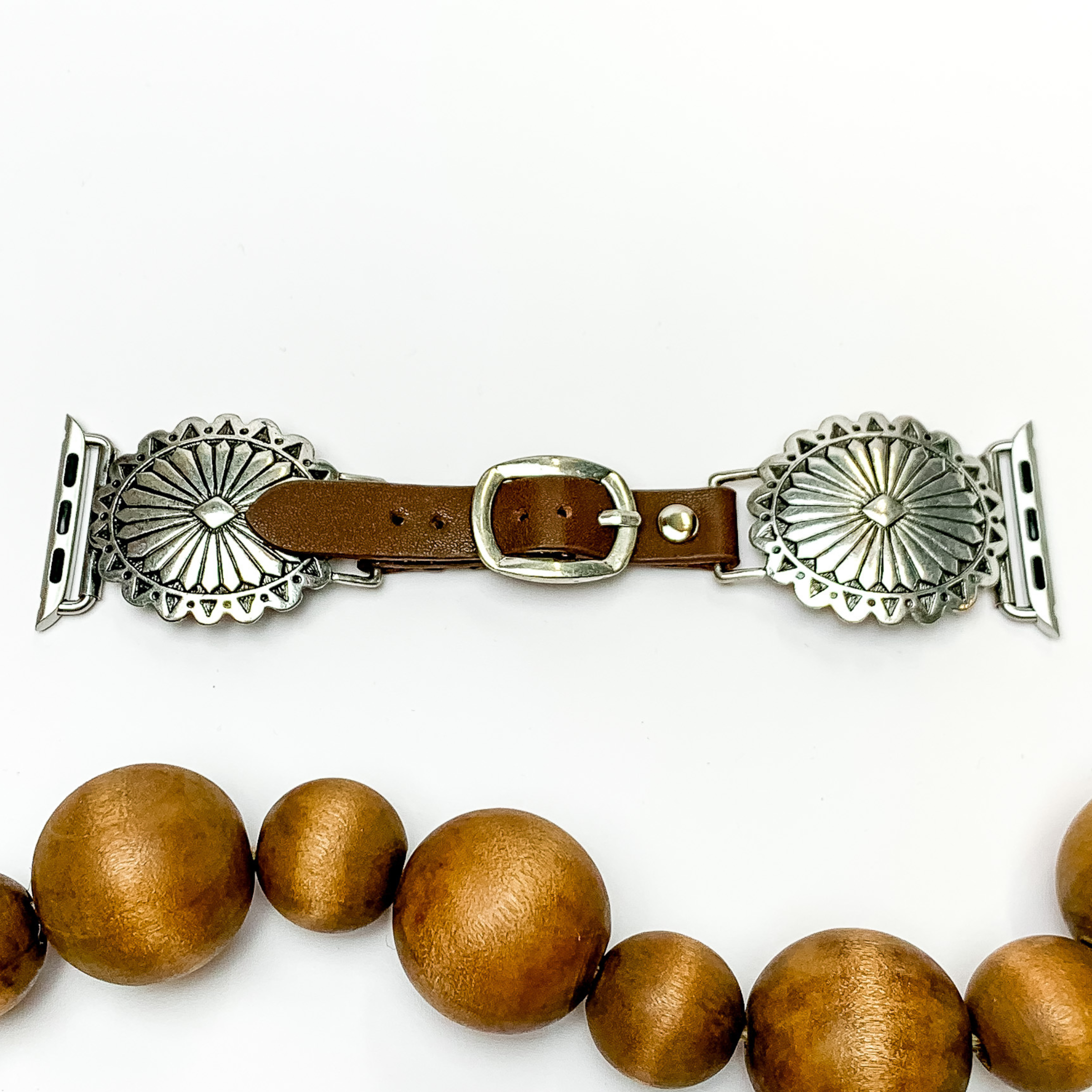 Dark Brown watch band with silver, oval concho ends with Apple watch band acessories. The conchos have engraved detailing. This watch band is pictured on a white background with brown beads underneath the band. 