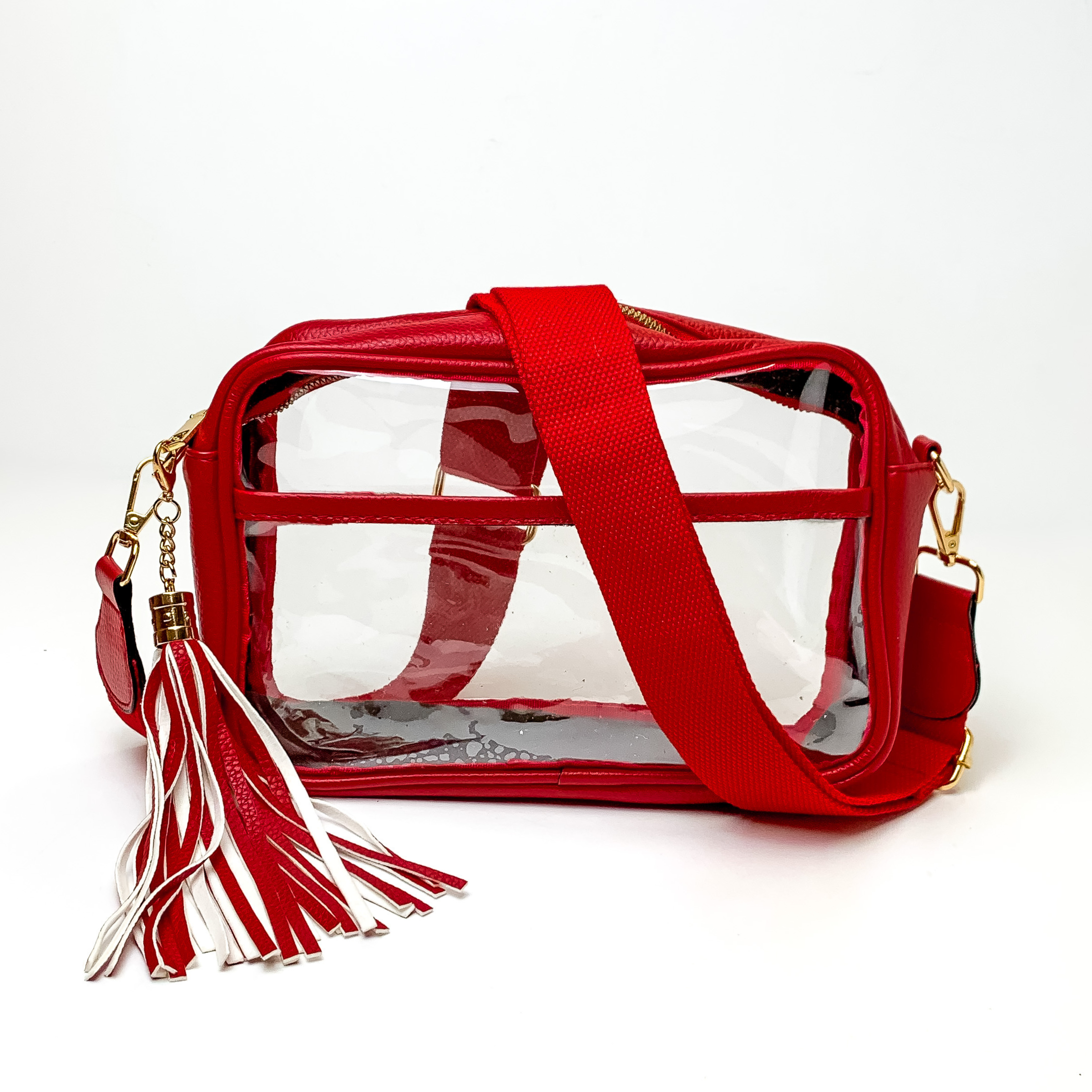 Pictured on a white background is a clear, rectangle purse with a red outline. This purse includes gold accessories, a red strap that is draped over the purse, and a red and white fringe tassel. 