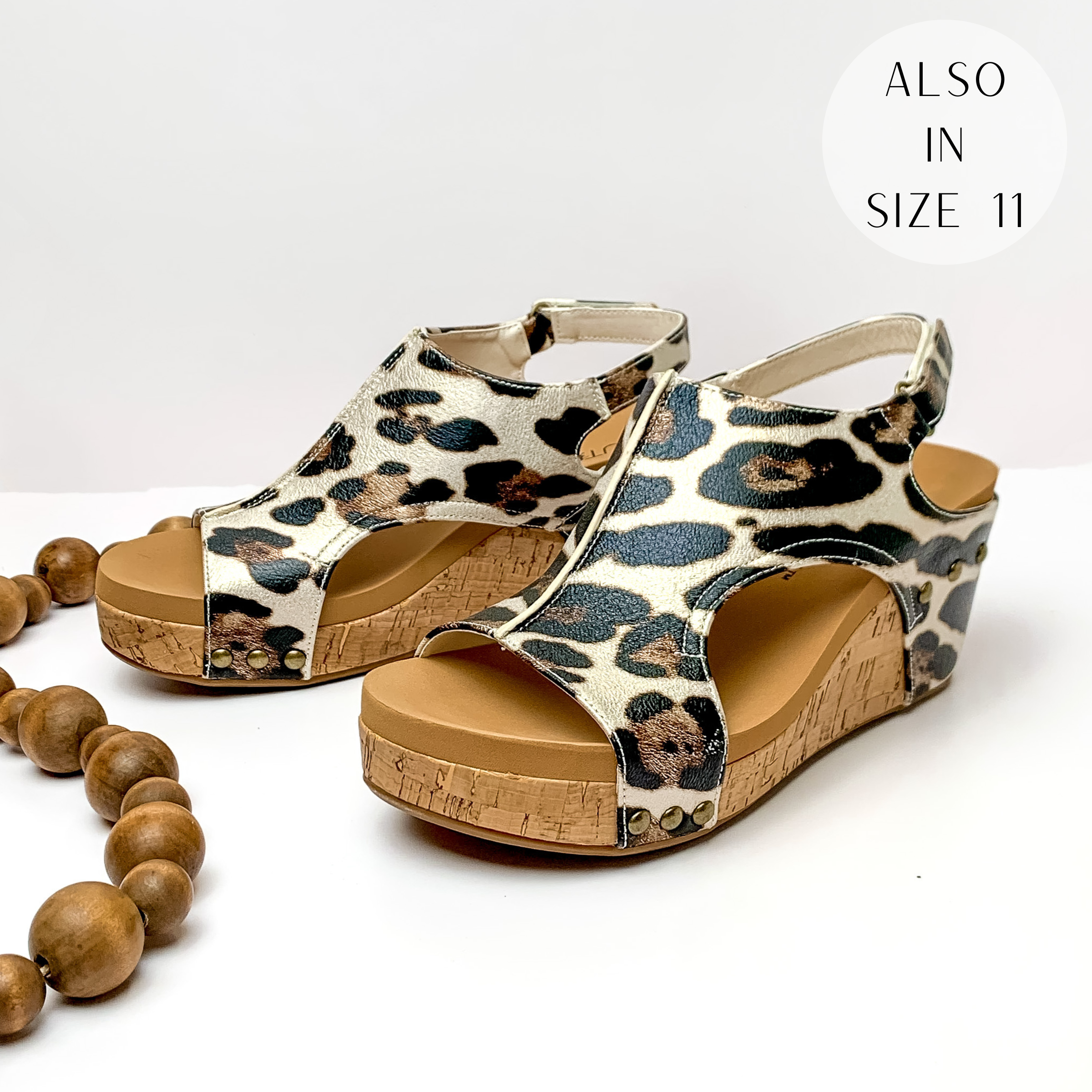Cork wedges with a ivory color upper with a black and brown leopard print and bronze studs connecting the upper to the wedge. These wedges also include a velco strap. These wedges are pictured on a white background with brown beads on the left side of the wedges.