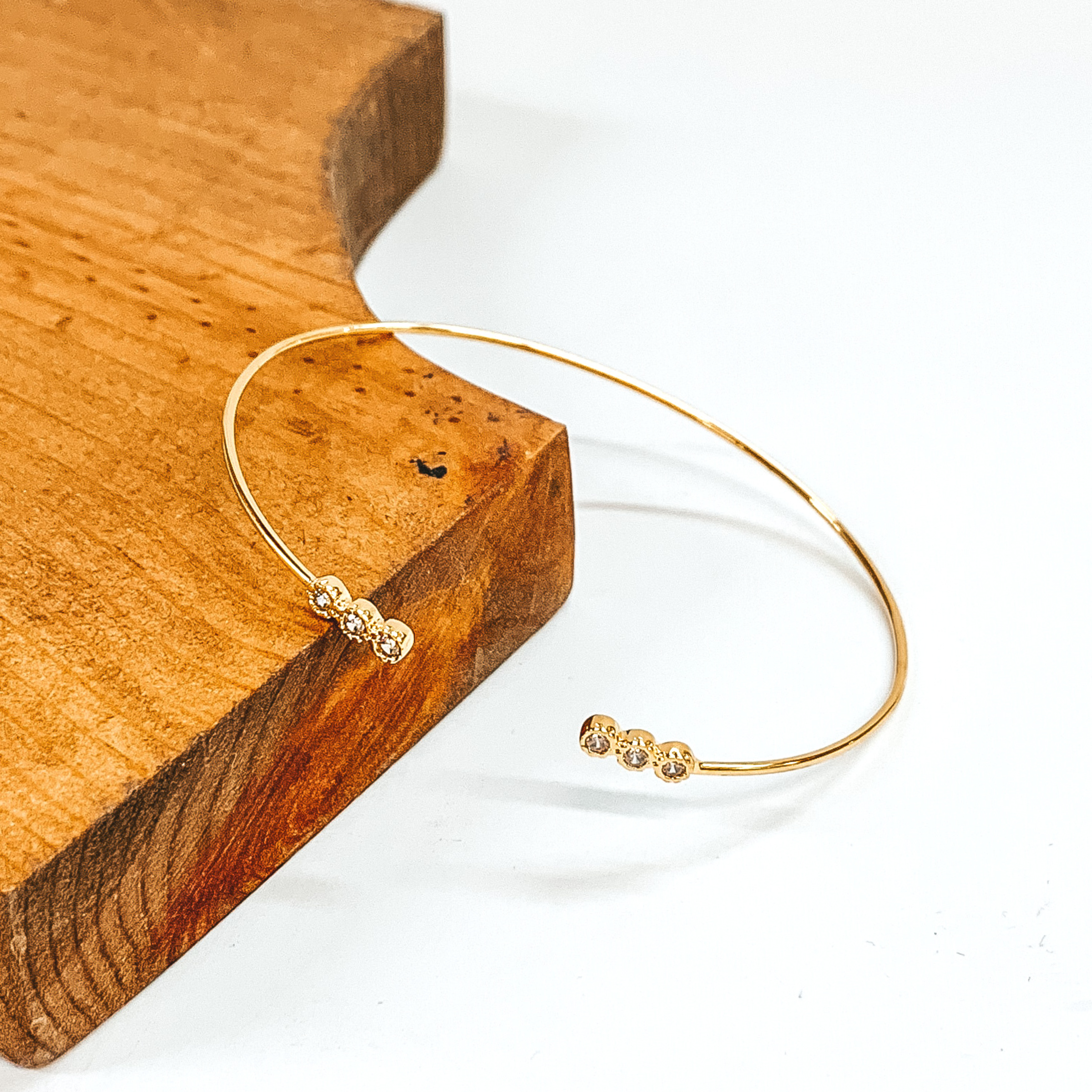 Thin, gold wire bangle with small, round tiny crystal ends outlined in gold. This bangle is pictured laying partially on a brown block on a white background. 