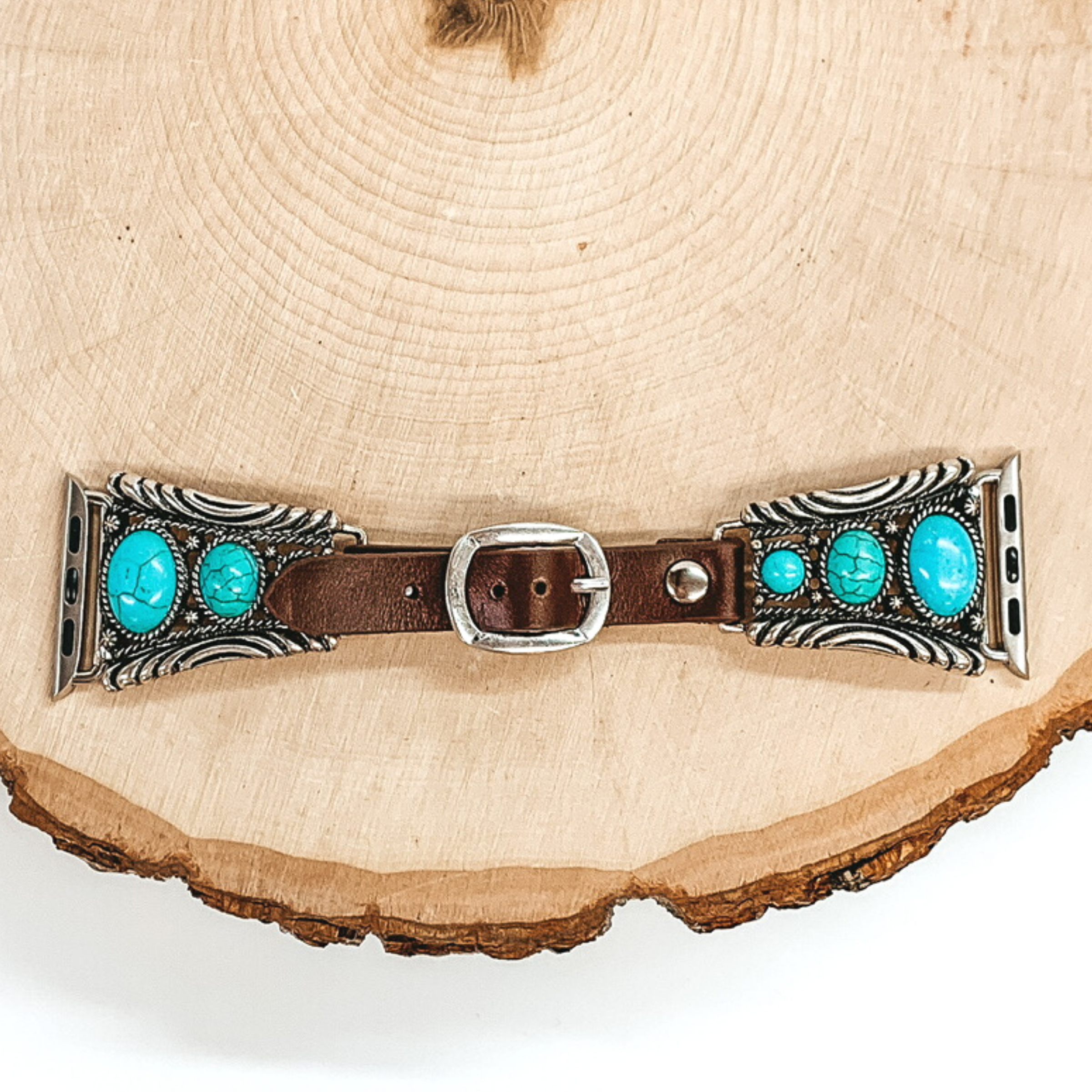 Dark Brown watch band with silver, trapesoid shaped ends with Apple watch band acessories. The ends have three turquoise stones each going from biggest to smallest. This watch band is pictured on a piece of wood on a white background.