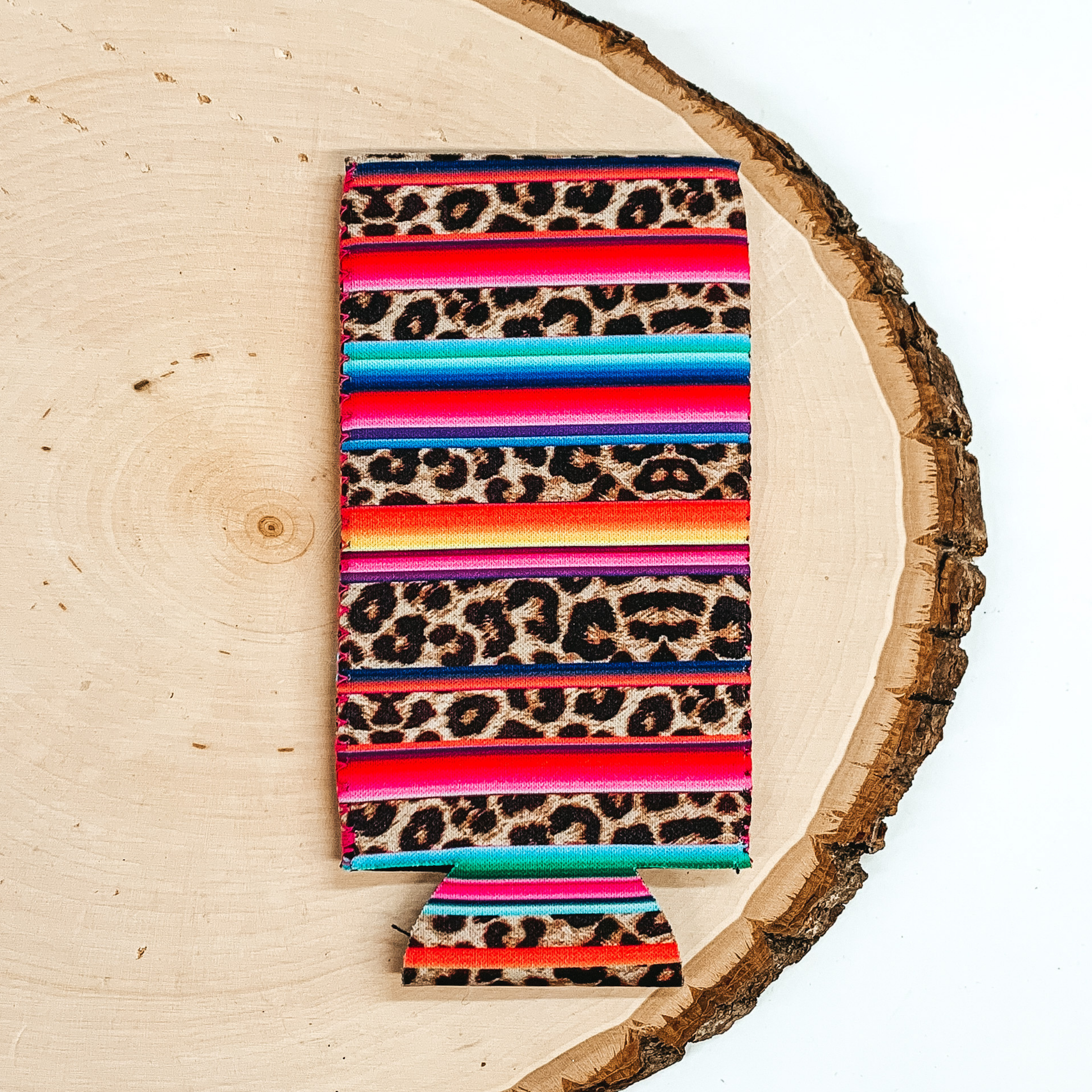 Striped serape and striped leopard print slim can koozie pictured laying on a piece of wood on a white background.