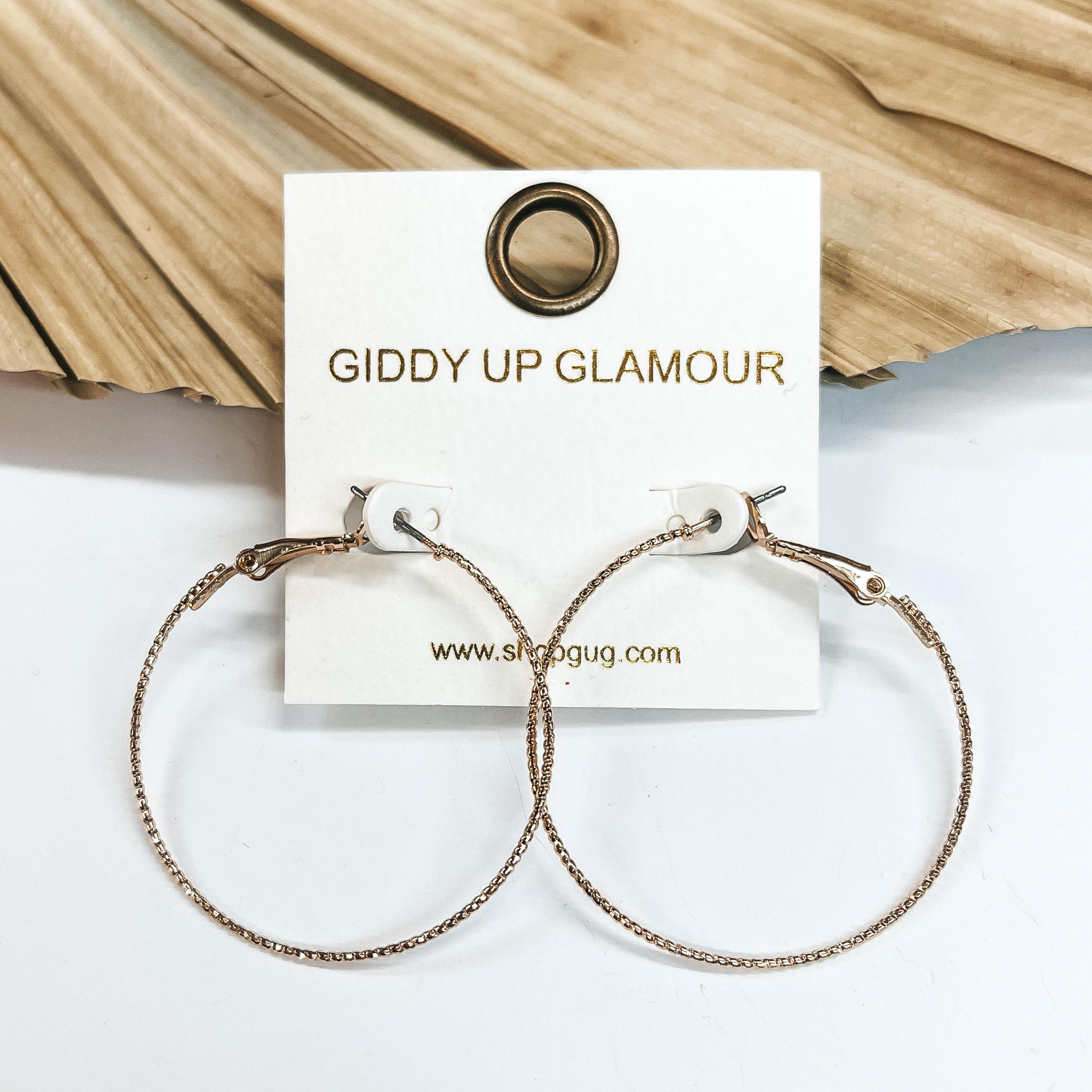 Thin wired gold hoops with rope texture. Taken on a white background, leaned up against a  dried up palm leaf.