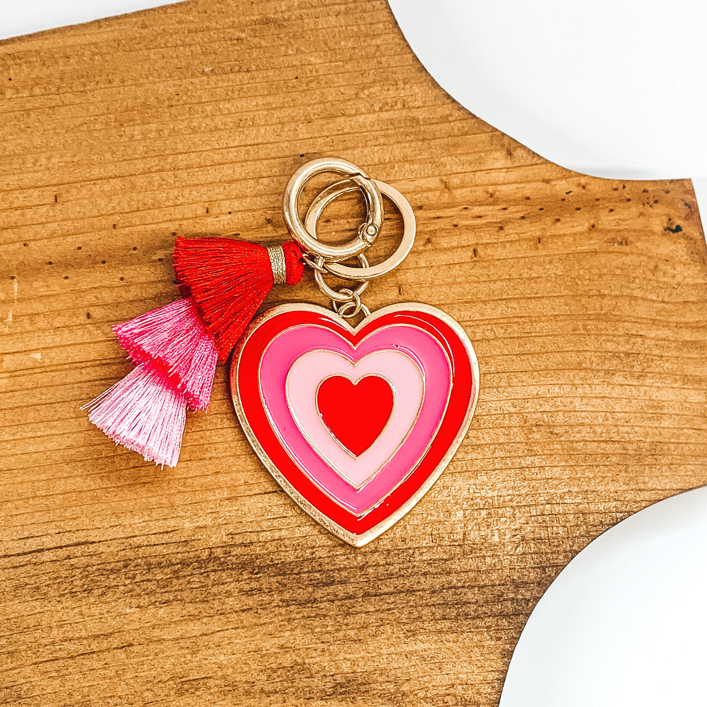 Gold heart pendant key chain with a tassel in that include the colors red, pink, and light pink. One side of the heart includes layers of hearts that includes the colors red, pink, and light pink. The other side is gold with an engraved outline in black and "xoxo' engraved in black. This key chain is pictured laying on a tan block on a white background. 