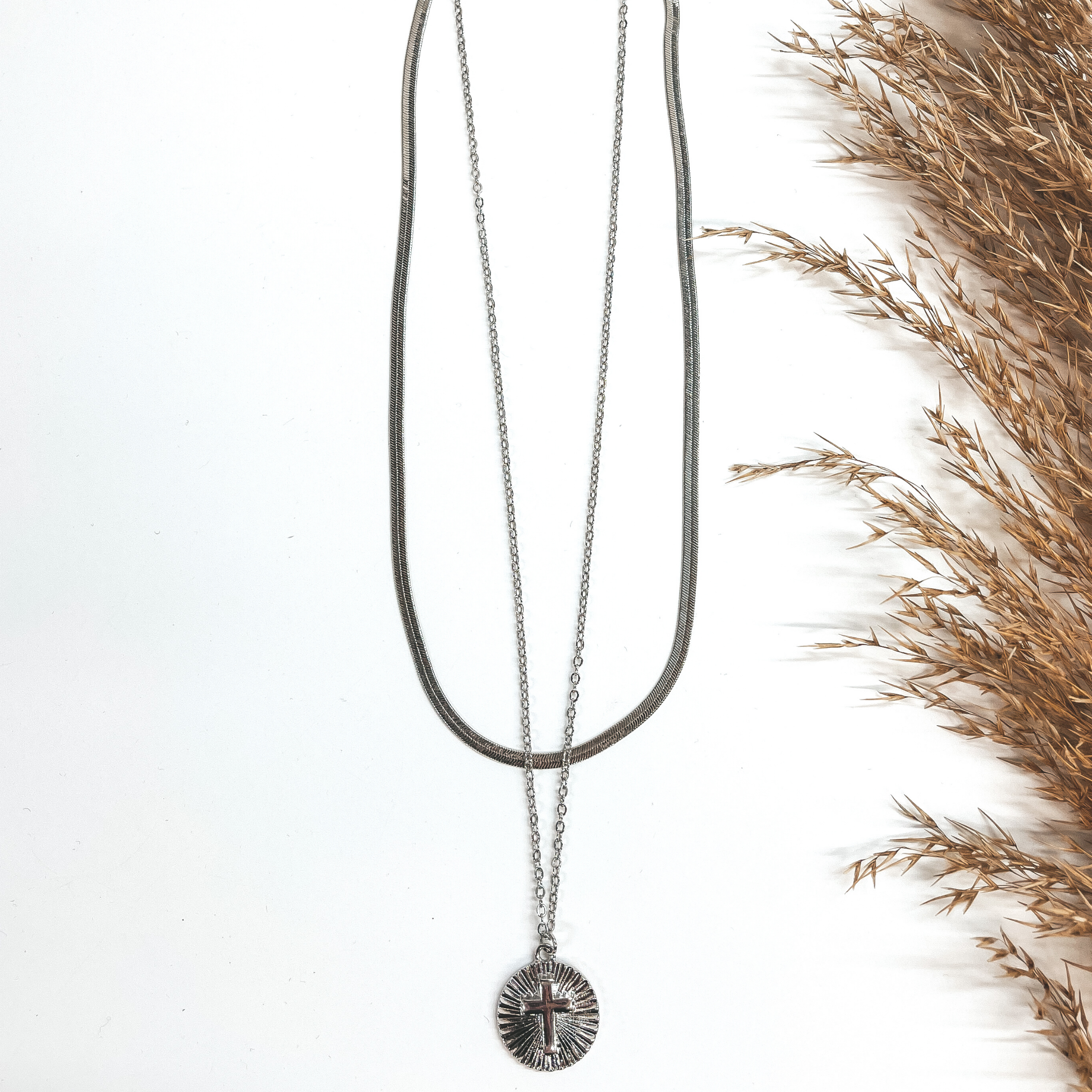 This is a double chained necklace in silver with a  circle pendant. The shorter chain is a snake chain,  the longer chain has a circle sunburst pendant with  a cross. This is taken on a white background and a  brown plant in the side.