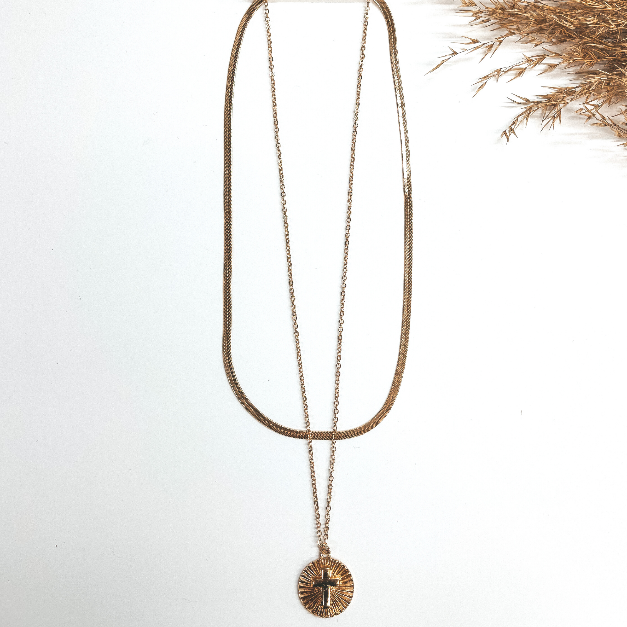 This is a double chained necklace in gold with a  circle pendant. The shorter chain is a snake chain,  the longer chain has a circle sunburst pendant with  a cross. This is taken on a white background and a  brown plant in the side.
