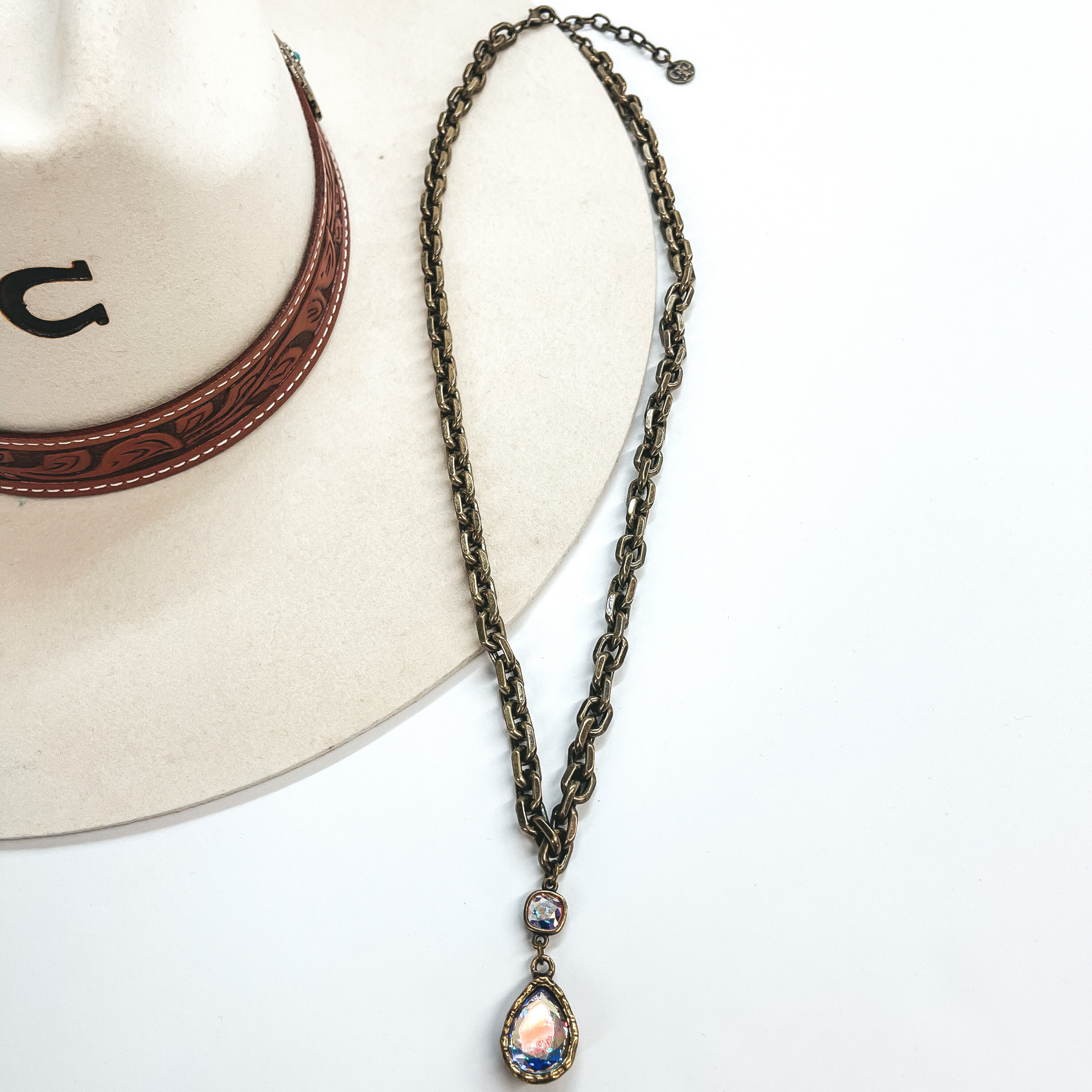 This is a long, thick link chain necklace in bronze  with an AB cushion cut crystal connected to an AB  crystal teardrop. This necklace is taken laying on  an ivory colored Charlie Horse hat and white  background.