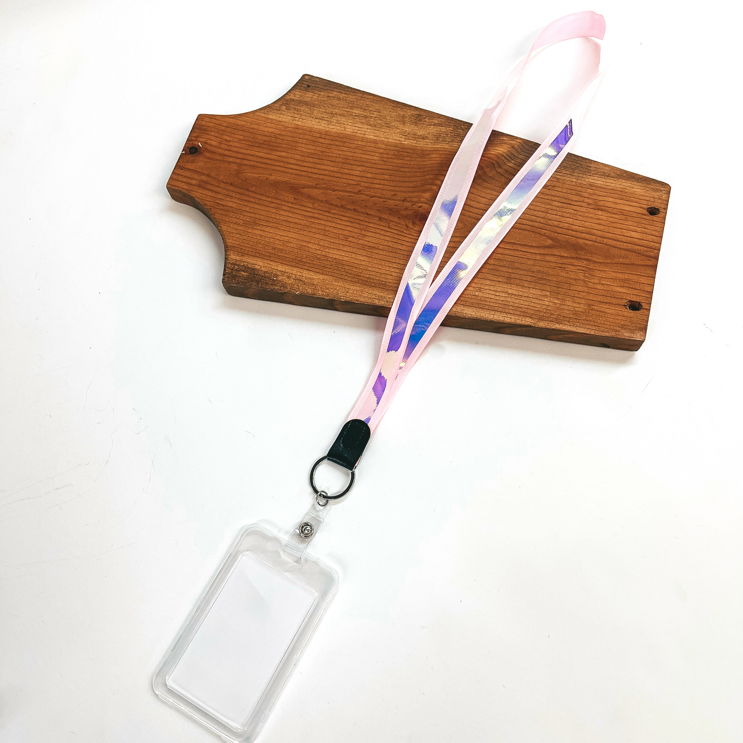 Pink ribbon lanyard with holographic center and clear ID holder, this  lanyard is laying on a brown necklace holder and on a white background.
