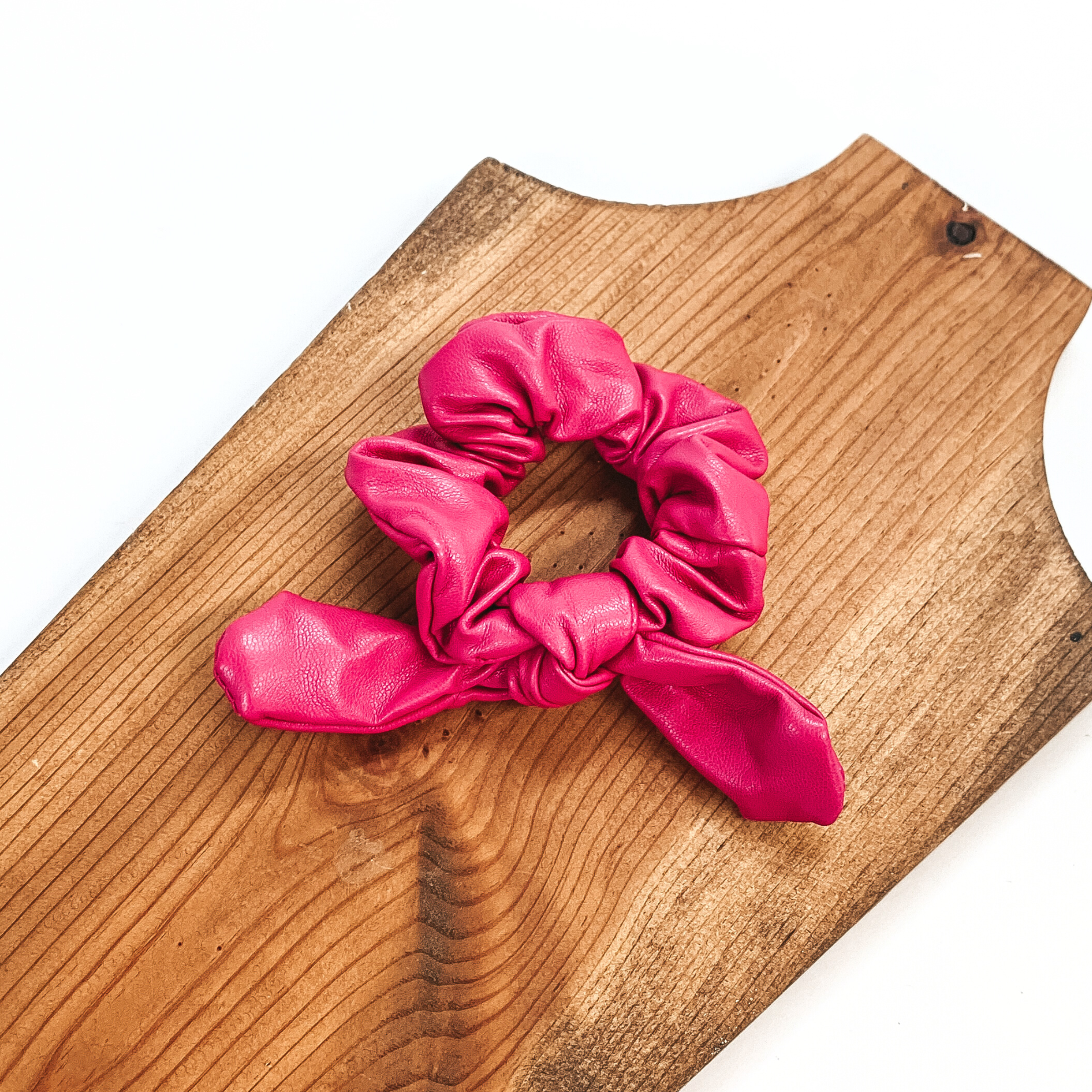 This is pink faux leather scrunchie with a bow, this scrunchie is taken on  a brown necklace board and on a white background.